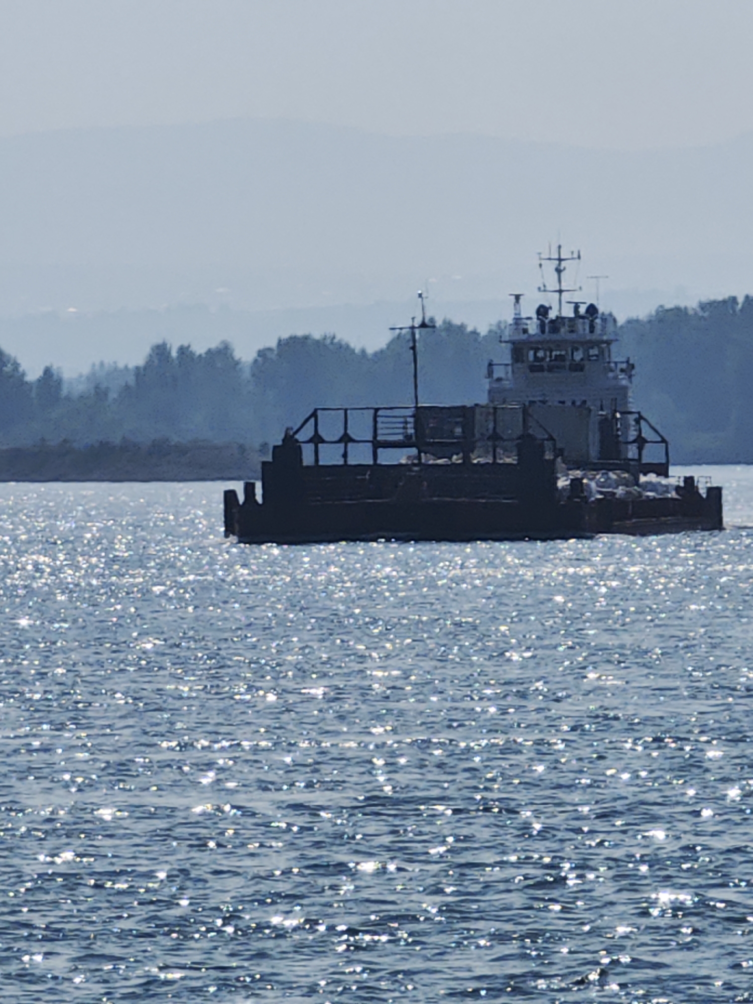 A barge with a superstructure is floating on the Yenisei River in Krasnoyarsk, Russia. The water is choppy and reflects sunlight, creating a glittering effect. The barge appears to be loaded with cargo or equipment. In the background, a hazy shoreline with trees and distant mountains is visible. The sky is overcast and pale blue. The overall atmosphere is serene and captures a moment of riverine activity.
