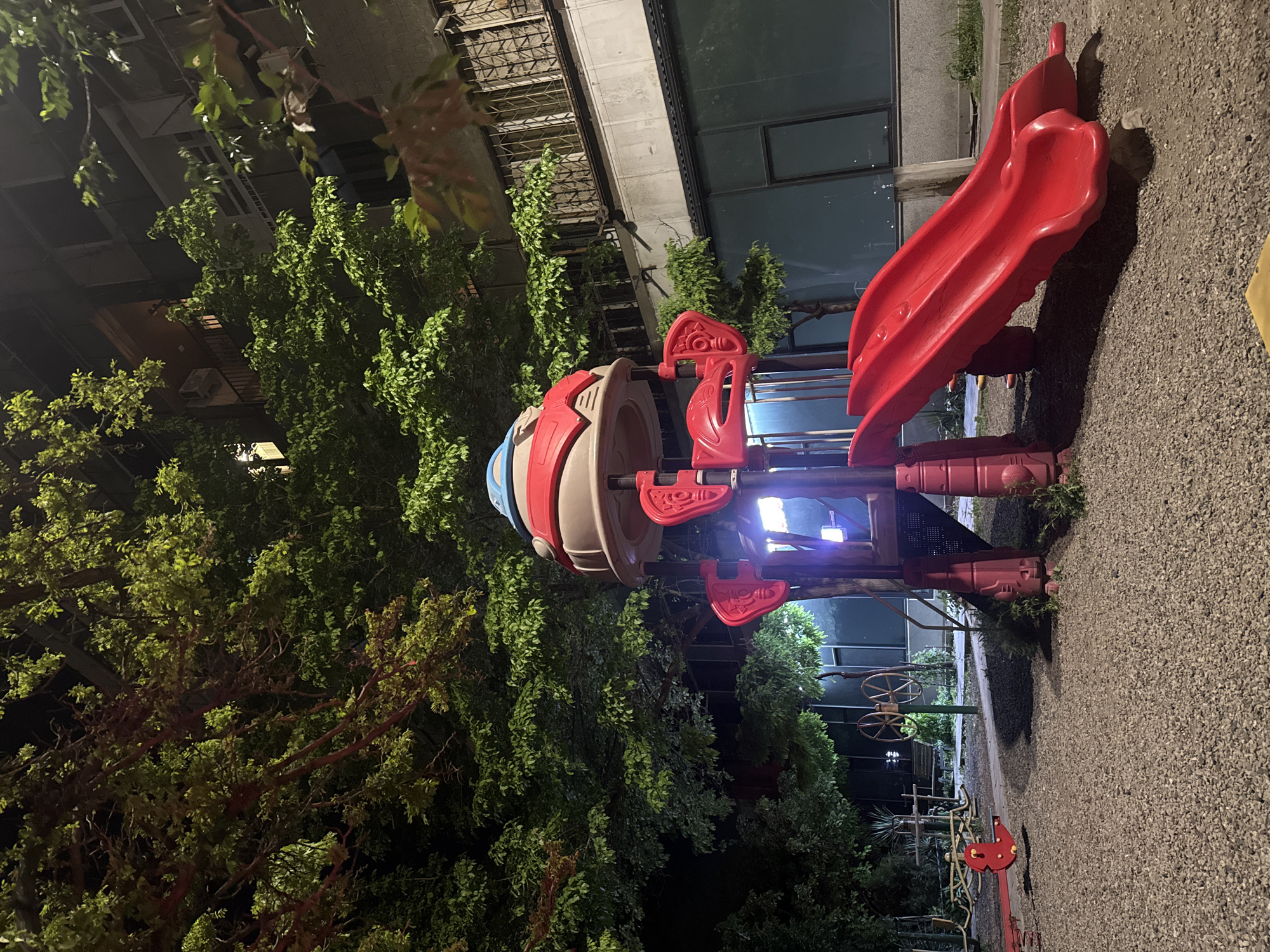 The image shows a playground at night, likely in Yerevan, Armenia. The main subject is a red and tan playground structure with a rocket ship design and a slide. It stands on a gravel surface surrounded by lush green trees and foliage. In the background, a building with what appears to be a staircase or metal railing can be seen, along with other playground equipment. The scene is illuminated by artificial lighting, casting shadows and highlighting the playground equipment. The image appears to be taken from a low angle, emphasizing the playground structure. There are no people visible in the picture.