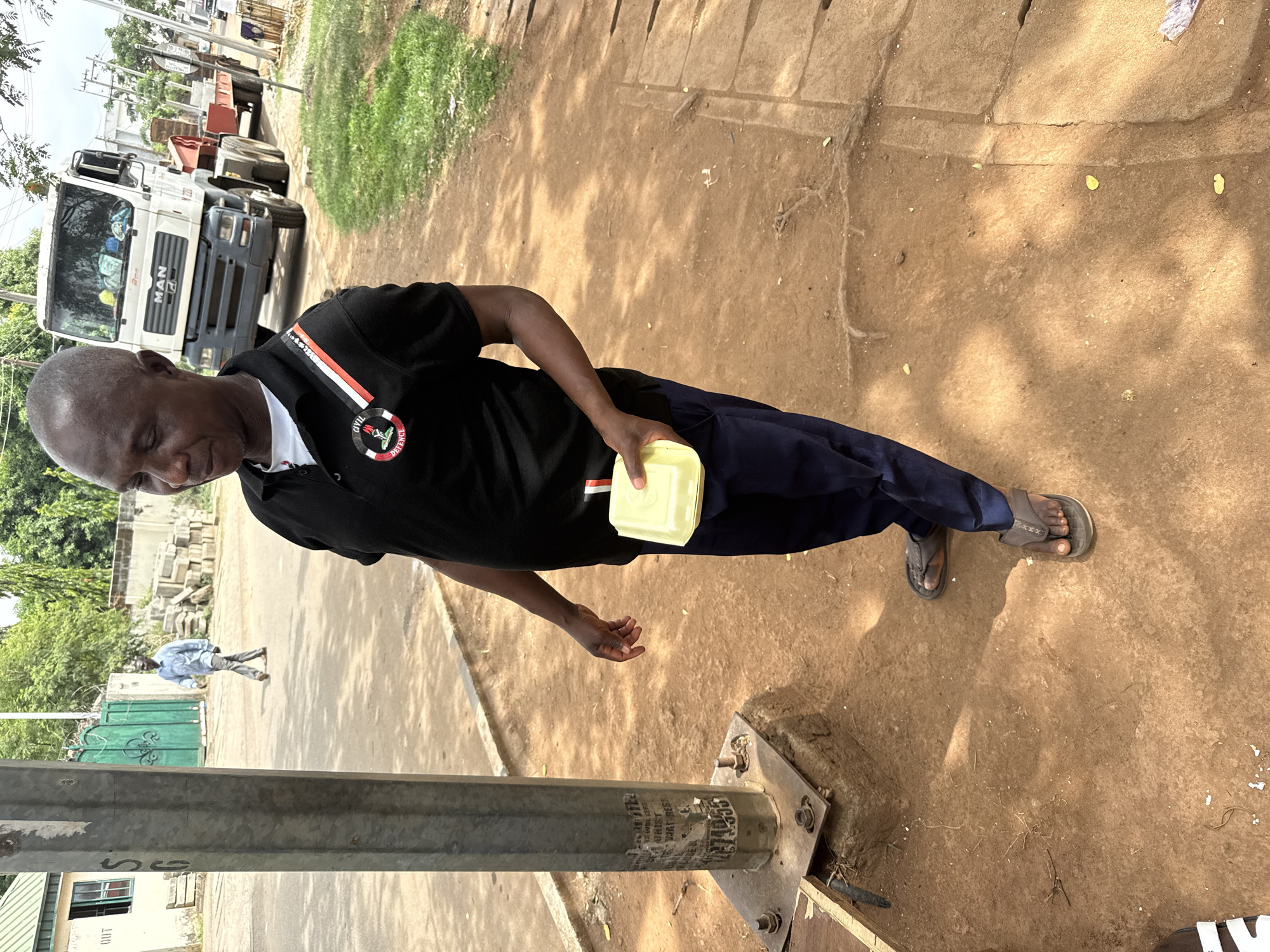 A man stands on a dirt and concrete surface, holding a yellow rectangular container. He is wearing a black t-shirt with a Civil Defence emblem, dark blue pants, and sandals. A truck is parked on the side of the road in the background. A man walks away in the background, and a light post is in the foreground. Based on visual details, the scene appears to be in an urban area, possibly in Abuja, Nigeria, as suggested by the request.