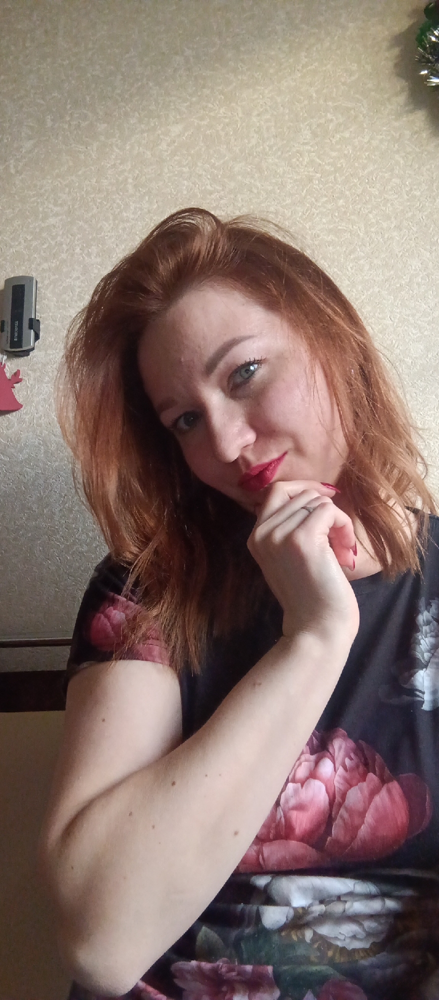 A woman with auburn hair, light skin, and red lipstick is posing indoors, resting her chin on her hand. She is wearing a floral print top with large pink flowers. A telephone or intercom system is affixed to the wall behind her, along with a small red decorative item. The wall itself is beige with a patterned texture. A green and silver tinsel decoration is visible in the upper right corner. The location is an indoor setting. The city is Unknown, Unknown.
