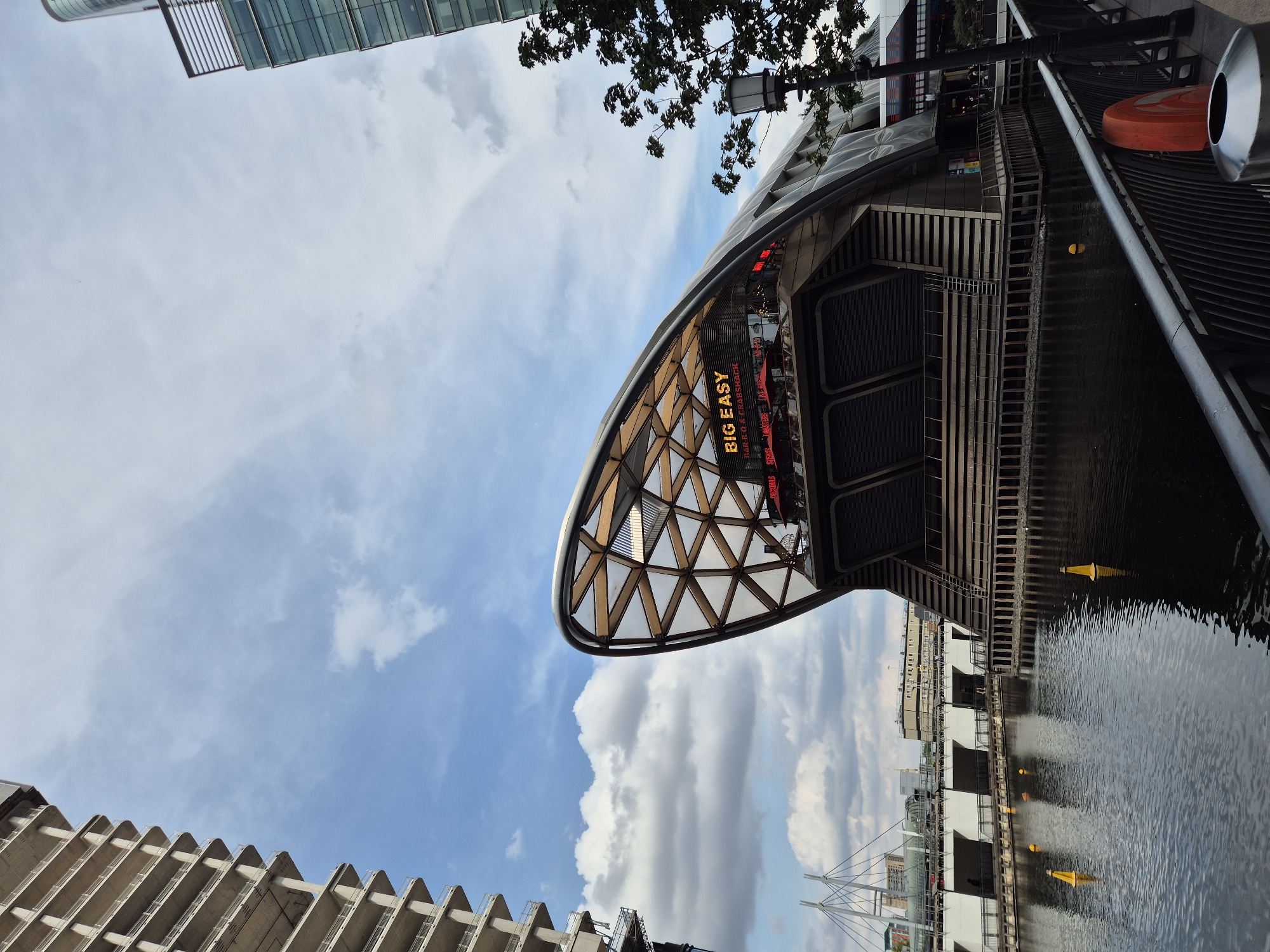 The image shows a futuristic building with a curved, shell-like roof structure made of wood and glass. The building, identified by a red sign as "BIG EASY BAR.B.Q & CRABSHACK," appears to be a restaurant or entertainment venue. It is situated on the waterfront, with calm water reflecting the sky and the building. In the background, there are other tall buildings, a bridge with cables, and a cloudy blue sky. The scene suggests an urban environment, likely a modern development area within the city of London, United Kingdom, possibly Canary Wharf given the architectural style and waterfront setting. There are no visible people in the foreground or midground. A black lamppost stands on the right side of the frame, and a metal railing runs along the edge of the waterfront.