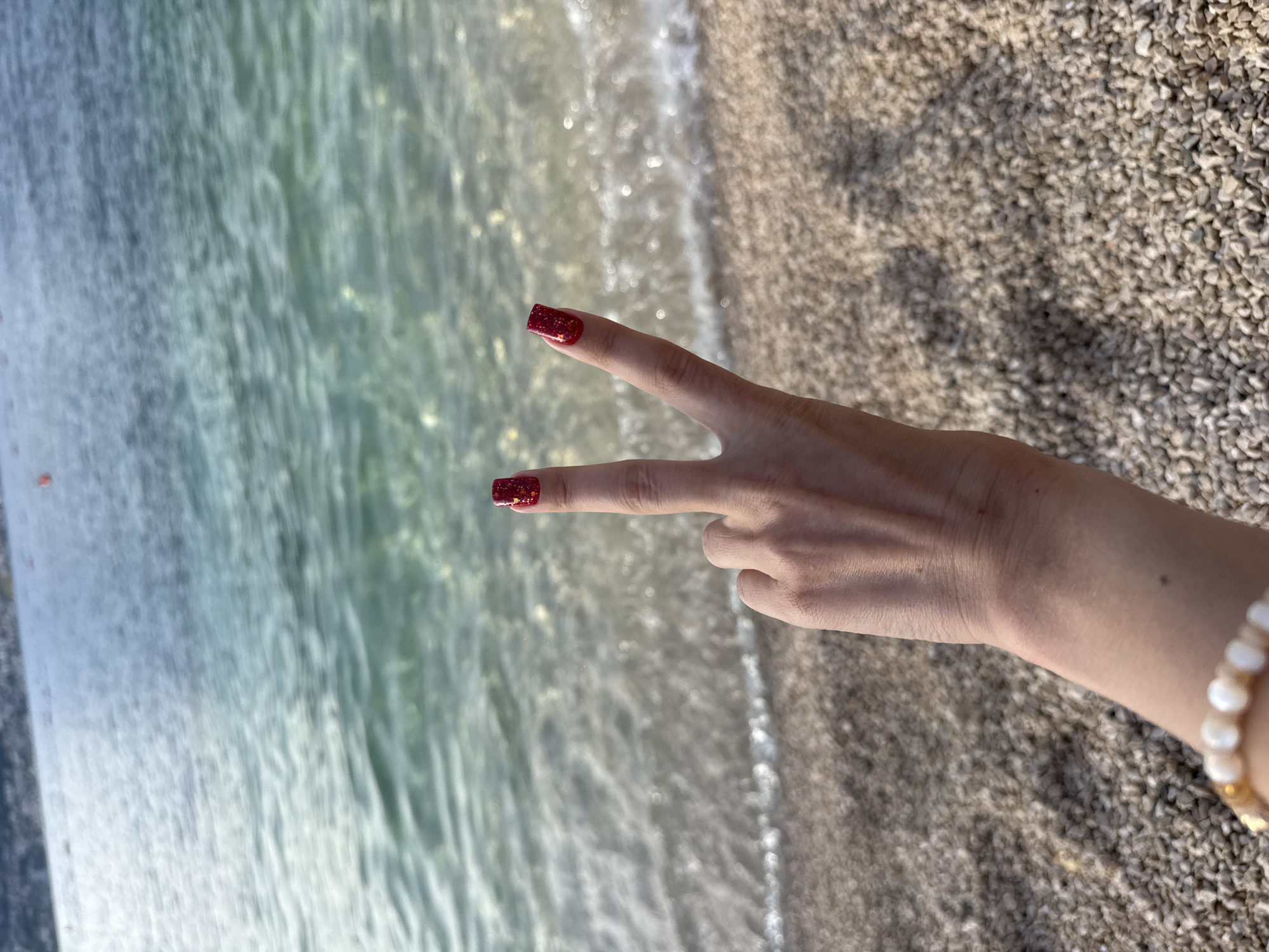 A close-up, high-angle shot captures a person's right hand forming a peace sign (V-sign) against the backdrop of a pebbled beach and the sea. The hand, positioned in the lower-center, has fair skin. The index and middle fingernails are adorned with square-shaped, glittery red nail polish containing small golden flecks. A light-colored beaded bracelet, composed of white and gold spherical beads, is visible on the wrist at the bottom left of the frame.
The foreground consists of tightly packed, light-colored pebbles forming the beach. Beyond the hand, the clear, shallow water of the sea gently washes onto the shore, creating white foamy lines. The water transitions from a light turquoise-green near the shore to a deeper blue further out, revealing the pebbled seabed beneath its surface. In the distant background, a blurred landmass or mountain range is visible on the horizon, with several small, red, indistinct objects floating on the water's surface, possibly buoys. The overall lighting suggests daylight.