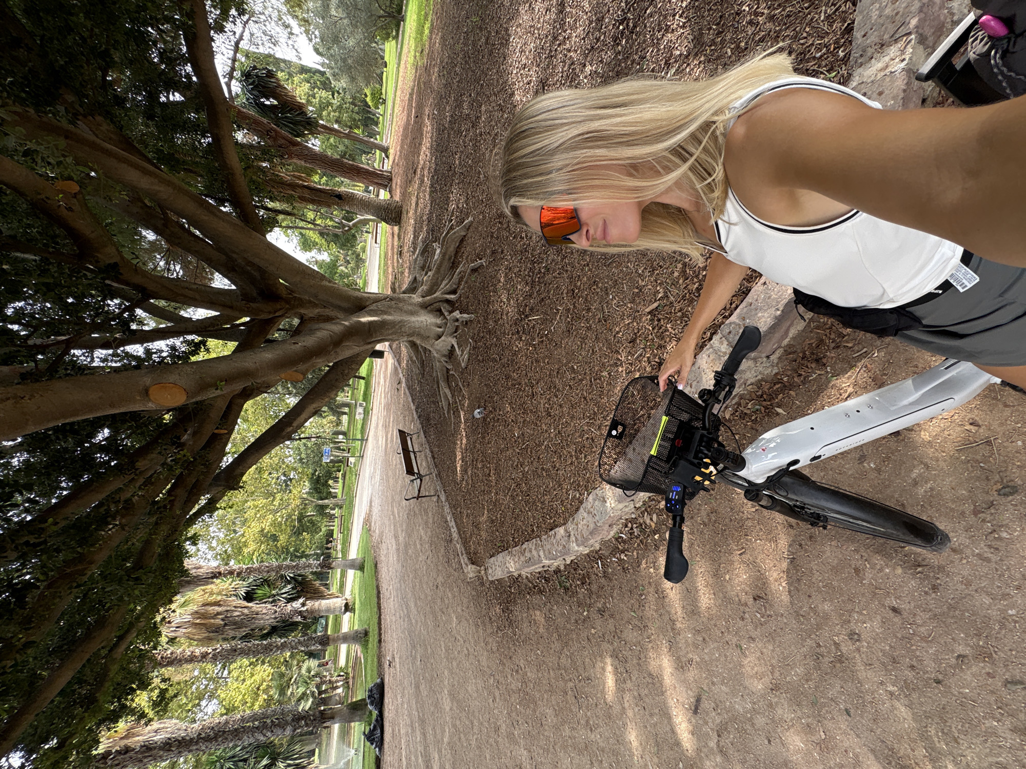 In the Jardí del Turia park in Valencia, Spain, a blonde woman with orange-tinted sunglasses takes a selfie next to a white electric bicycle. The woman wears a white tank top with black trim and dark shorts. She is positioned to the right of the image, facing left, with the bicycle handlebars and front basket in the foreground. The background shows a large, mature tree with exposed roots, a dirt path, palm trees, and other lush greenery. The ground is covered in mulch and fallen leaves.