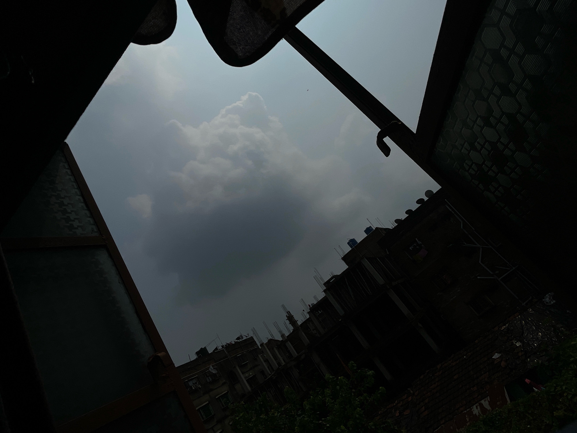 This image captures a view of the sky and buildings in Bhatpara, India, as seen from an interior perspective looking outward. The foreground is dominated by dark, silhouetted structures, possibly a window frame and some sort of awning or covering, which create a triangular opening. Through this opening, a cloudy sky is visible. A large, fluffy white cloud stands out against a backdrop of a darker, overcast sky. Below the sky and clouds, the tops of several buildings are visible. These appear to be multi-story structures, some of which seem to be under construction with exposed rebar. The overall lighting suggests it may be dusk or an overcast day. The buildings are characteristic of an urban environment, with visible windows and some clutter on rooftops. The scene is tilted, as if the camera or the viewpoint is angled.