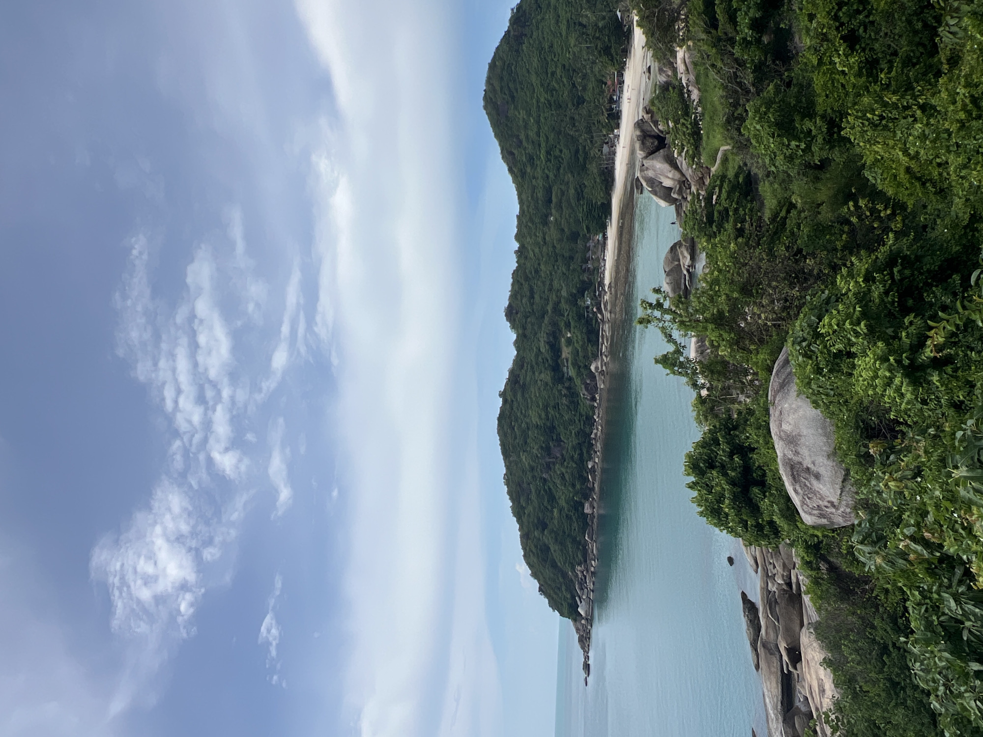 The image depicts a coastal scene overlooking a bay in Baan Chaweng Noi, Thailand. The water is a calm, turquoise blue, meeting a sandy beach that curves into the distance. To the left of the bay, a rocky outcrop juts into the sea, covered in lush green vegetation. Behind the beach, a densely forested hillside rises, its slopes covered in trees. The sky above is a pale blue, with scattered white clouds. In the foreground, large grey boulders are interspersed with dense green foliage. A few distant figures are visible on the beach, appearing as small specks against the expanse of sand. The overall impression is one of a serene tropical landscape.