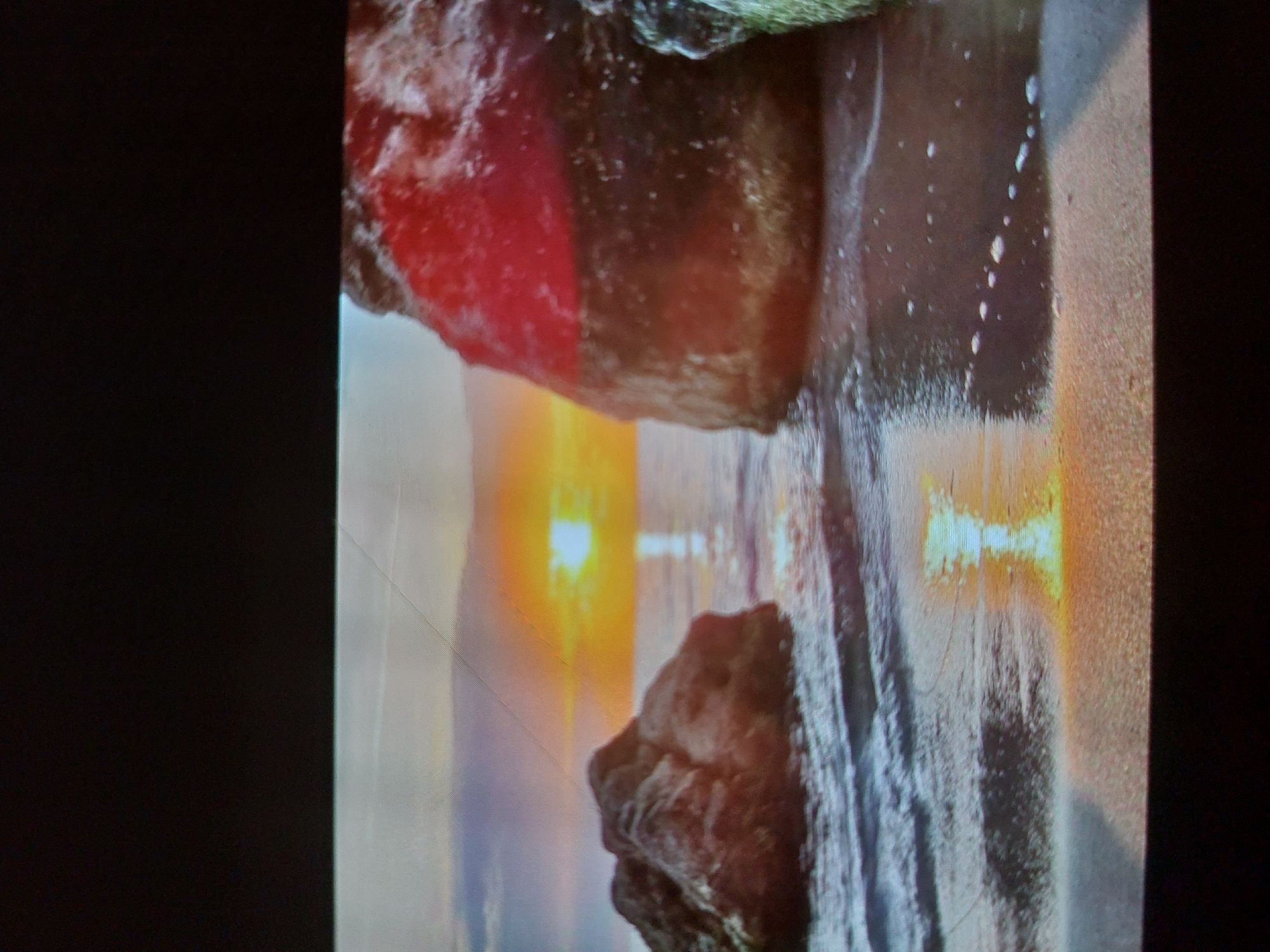 The image, appearing to be a photograph of a display screen, depicts a vibrant sunset or sunrise over a calm body of water, likely a sea or ocean, in a landscape orientation. A bright, low-hanging sun dominates the horizon, casting an intense golden-orange and yellow light that reflects vividly on the water's surface, creating a shimmering path of illumination.
In the foreground, two large rocks frame the scene. The rock on the left (when viewed in its natural landscape orientation) is reddish-brown with rugged textures, while the rock on the right is darker and also textured. The water around the rocks shows subtle ripples and reflections of the sky's soft hues, which transition from bright orange and yellow near the sun to lighter blues and grays further up. Some white speckles, possibly sea spray or foam, are visible near the rocks and on the water.
The time of day is clearly dawn or dusk, characterized by the dramatic colors and low sun. The weather appears clear and calm. The scene suggests a coastal setting, consistent with Tsarevo, Bulgaria.
Visible details consistent with a photo of a screen include pixelation, subtle diagonal lines (possibly screen artifacts or scratches on the display), and black bars flanking the main image on the left and right, indicating the original content did not fill the entire frame. No people or text are visible.