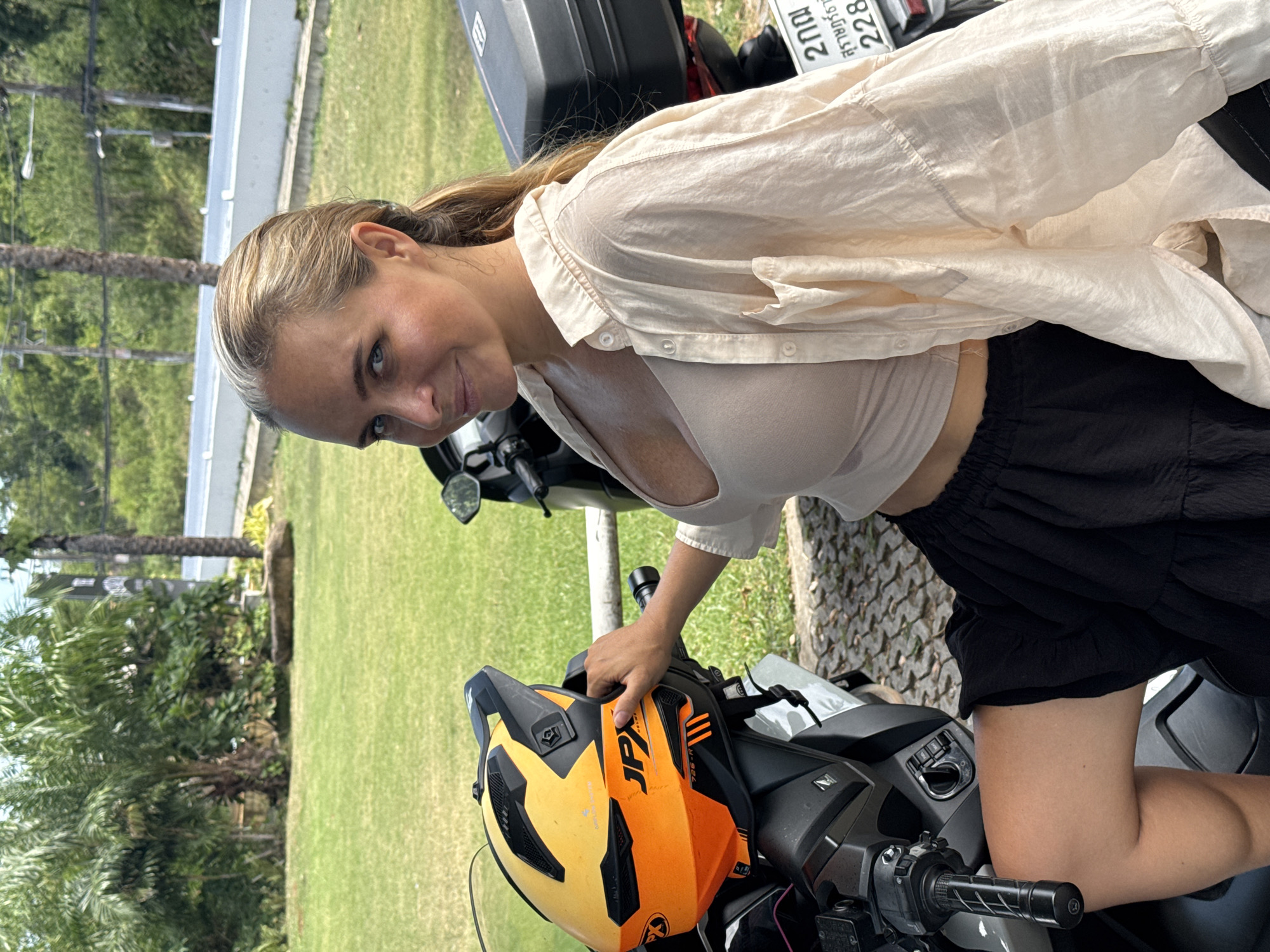 A woman with blonde hair, tied in a ponytail, sits on a scooter. She is wearing a light beige crop top and a loose, open beige button-down shirt. A black, ruffled skirt is visible. Her left hand rests on a yellow and orange motorcycle helmet. The scooter is parked on a cobblestone surface, with a grassy area behind it. Palm trees and other greenery are visible in the background, along with a white wall and what appears to be a road. A license plate with Thai characters is attached to the back of the scooter. The lighting suggests it is daytime.