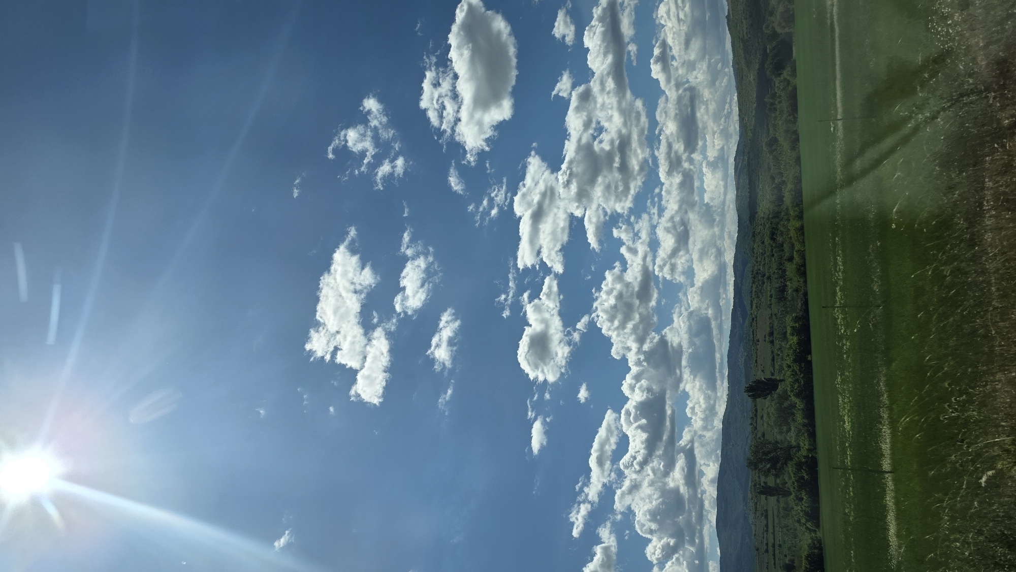 A landscape shot oriented with the ground on the right side of the frame and the sky on the left side of the frame. The bright sun shines from the bottom left corner of the frame. The sky is blue and contains scattered, white cumulus clouds. A line of mountains partially obscured by clouds runs diagonally across the middle of the frame. At the foot of the mountains is a dense green forest. The right side of the frame is filled with a green field. The scene is in Mursalevo, Bulgaria. There is no evidence of people in the image.
