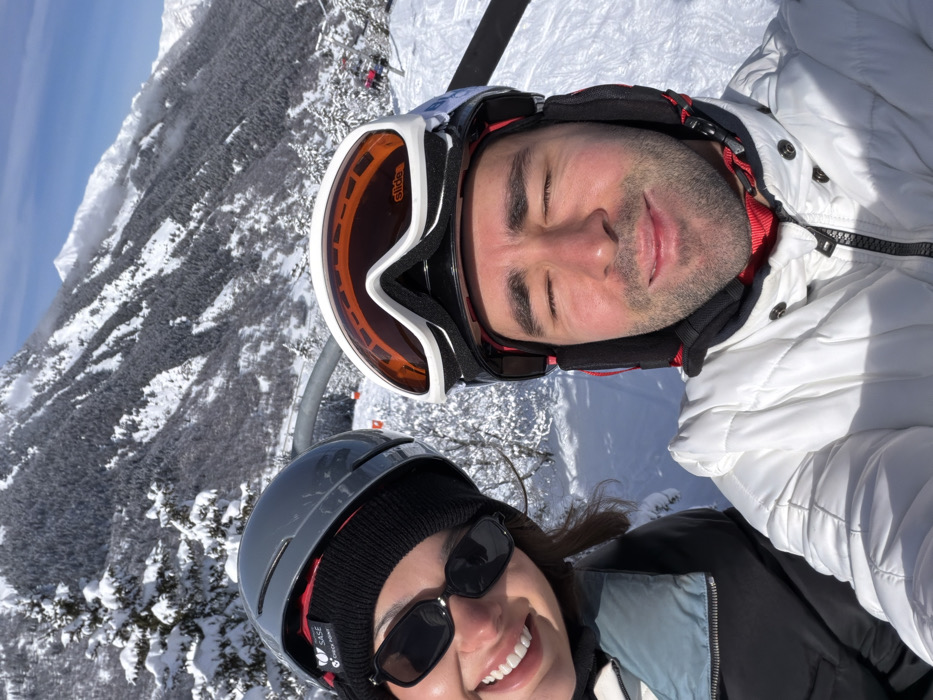 The image depicts two individuals on a ski lift in Les Allues, France, under bright daytime conditions. The female on the left, with dark hair, is smiling towards the camera. She wears a dark grey ski helmet with a "SASE CHECK POINT" sticker, a black beanie, black rectangular sunglasses, and a black and light blue ski jacket. The male on the right, with visible stubble, has his eyes closed and a slight smile. He is wearing a black ski helmet, with orange-tinted ski goggles pushed up onto the helmet, and a white quilted ski jacket with a dark collar featuring red and blue accents. They are positioned against a backdrop of steep, snow-covered mountains densely populated with snow-laden pine trees. A grey metal structure of the ski lift is visible behind them, along with a distant ski chair carrying other passengers on the slope below. Orange boundary markers are visible on the snow-covered terrain. The sky above is clear and blue with minimal cloud cover.