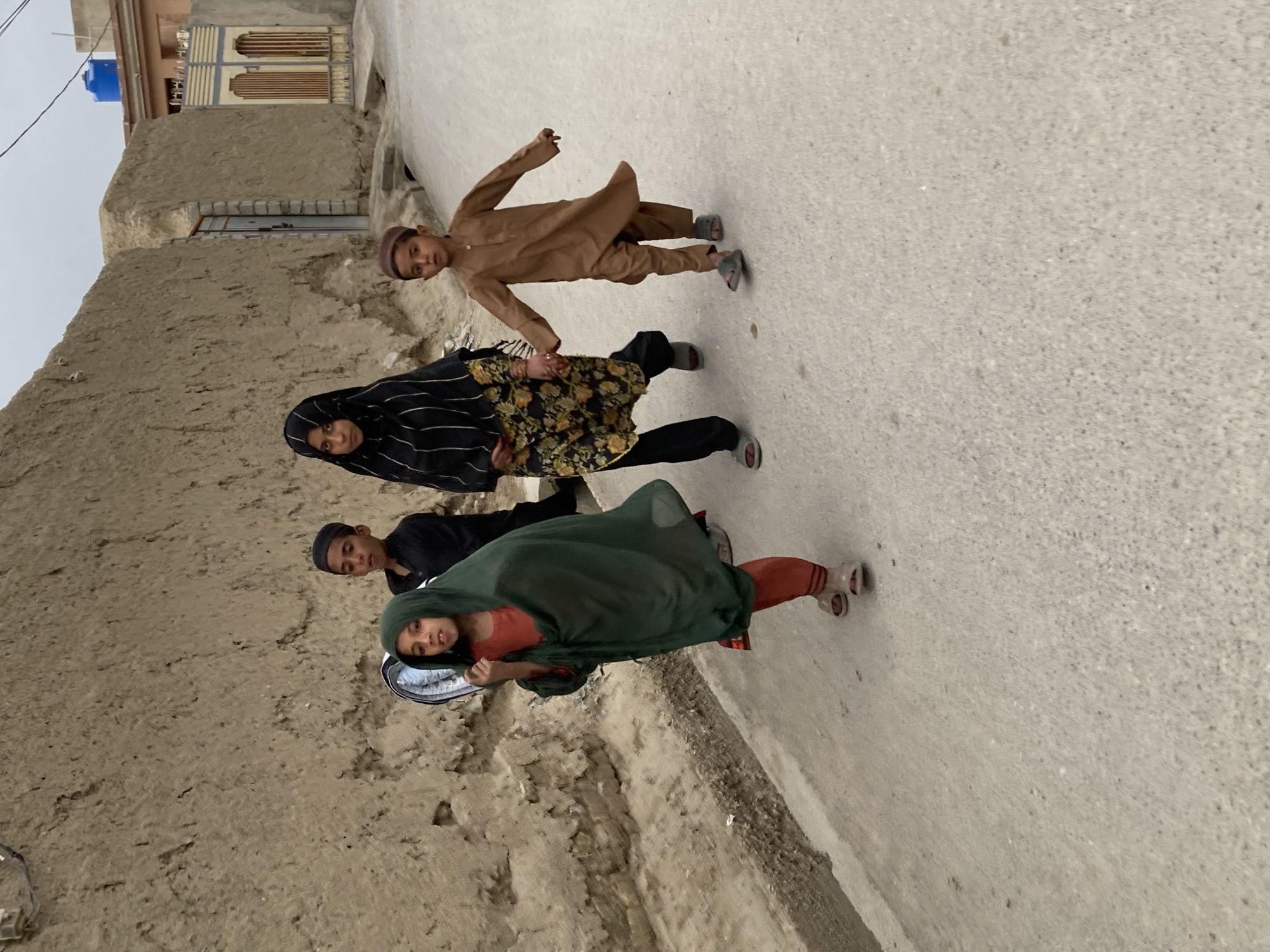 Four children are walking along a paved street in Quetta City Tehsil, Pakistan. The children are a mix of boys and girls. One child is wearing a brown outfit and a cap, another is in a black and floral outfit with a head covering. A third child wears black, and the fourth is covered in a green shawl. The buildings along the street are constructed of mud or plaster. The sky is overcast.