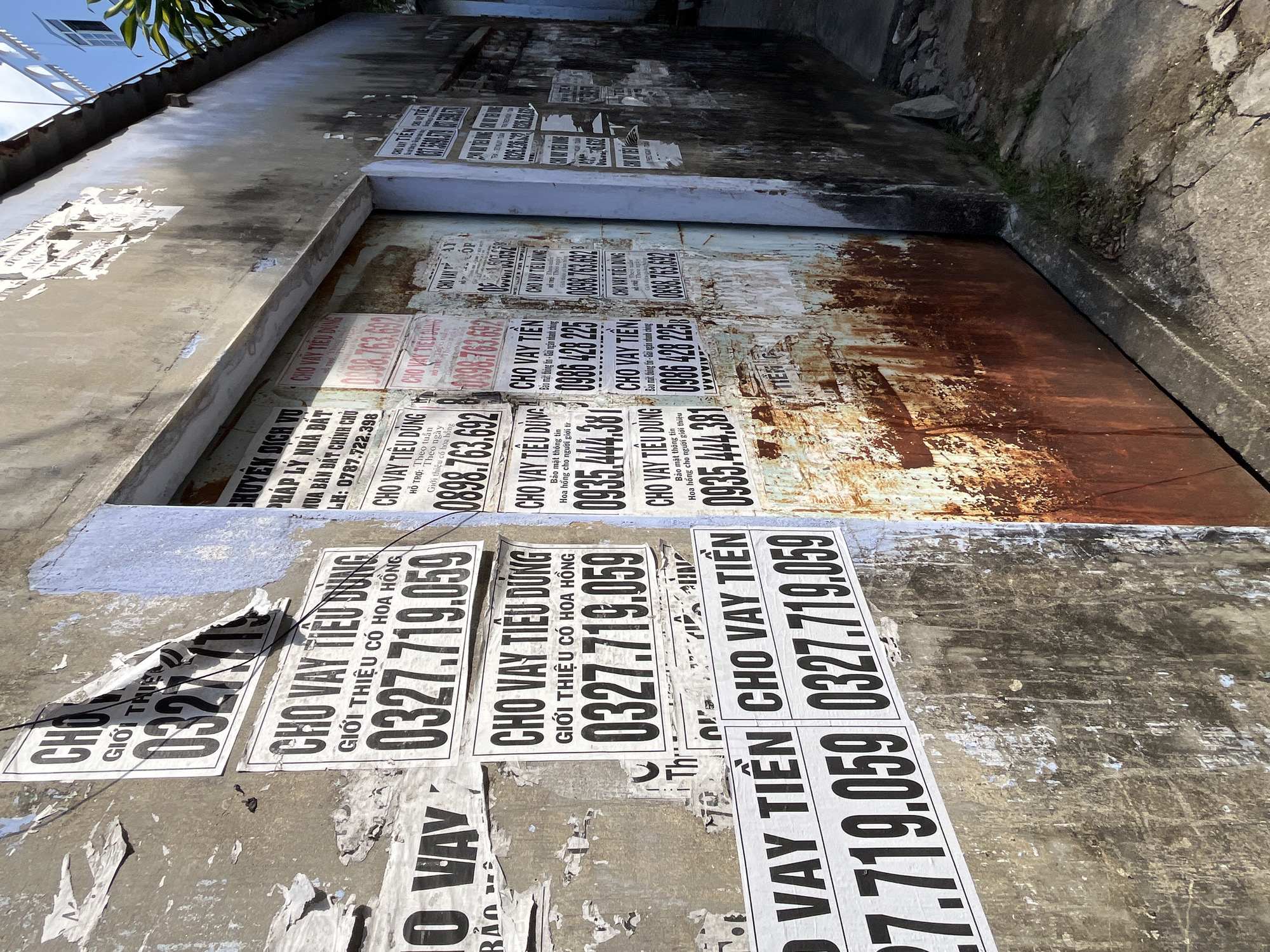 The image shows a narrow alleyway in what is presumed to be Bac Nha Trang Ward, Vietnam. The alley is flanked by two walls, one appearing to be part of a building and the other a natural stone wall. Both walls and a metal door-like structure are heavily plastered with commercial posters advertising loan services ("Cho Vay Tien" – Loan Money) and consumer loans ("Cho Vay Tieu Dung" – Consumer Loan). The posters display telephone numbers (e.g., 0327.719.059, 0898.763.692, 0935.444.381, 0988.428.225) and promotional phrases like "Gioi Thieu Co Hoa Hong" (Introduce to receive commission). The posters are in varying states of adhesion, with some torn and peeling. The metal structure has a rusty surface. The environment is urban and appears somewhat deteriorated. There are no people visible in the image.
