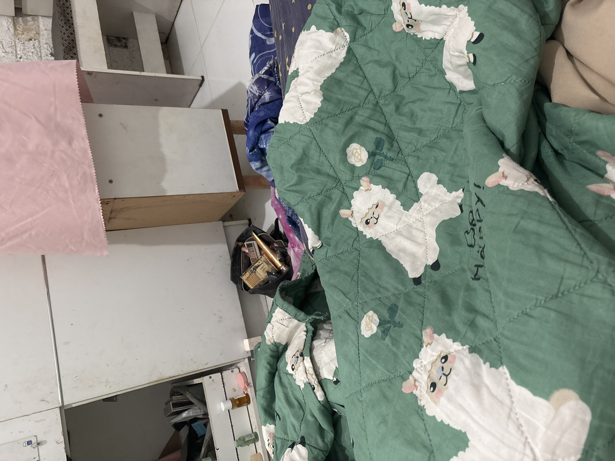 The image shows a messy bedroom scene in North Jakarta, Indonesia. The focus is on a green comforter with a repeating pattern of white cartoon alpacas and roses. Some of the alpacas have text that reads "Be Happy!". The comforter is draped over a bed, with other blankets and fabrics visible underneath. In the background, there is a white cabinet with a pink cloth draped over the top. The cabinet appears to be open on the left side, revealing some items inside, including what looks like a makeup bag or toiletry bag filled with various cosmetic items. The flooring is tiled.