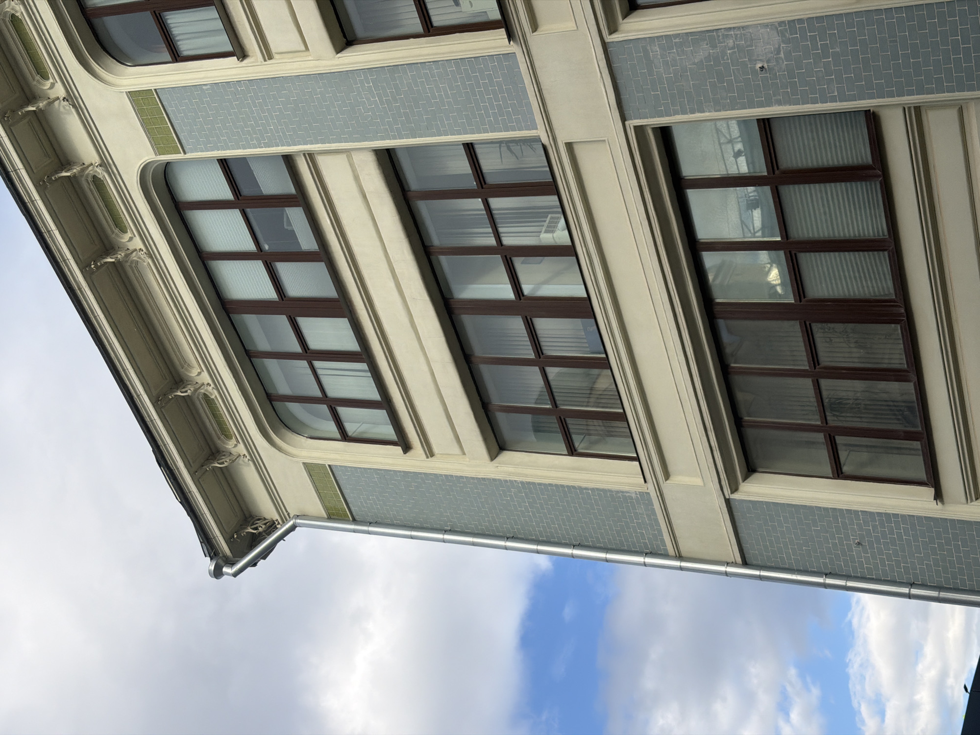 The image depicts a low-angle view of a building facade against a cloudy sky. The building features several rectangular windows with dark brown frames and multiple panes. The facade exhibits architectural details, including moldings, decorative cornices, and what appears to be light blue tilework or patterned masonry between the windows. A metal rain gutter runs along the edge of the building, juxtaposed against the sky. The building appears to be an older style with ornate details, but further identifying features of the location are not discernible. It is impossible to confirm Moscow as the location from this image.