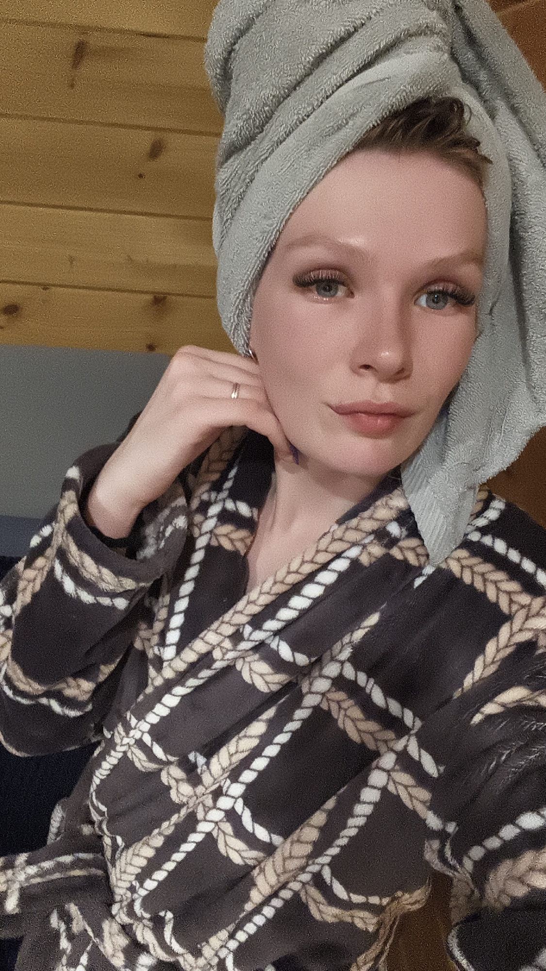 A woman with fair skin is captured in a close-up shot. She wears a light grey towel wrapped around her head and a brown and tan robe with a rope pattern. Her right hand is raised to her face, with her fingers gently touching her cheek. A simple ring adorns her finger. She has light blue eyes and appears to be wearing false eyelashes. The background is a wooden wall on the upper left and a grey wall behind her shoulder. The image might be taken indoors, possibly in a bathroom or bedroom. There is no information given about the location of the image in Русский Вожой, Russia.