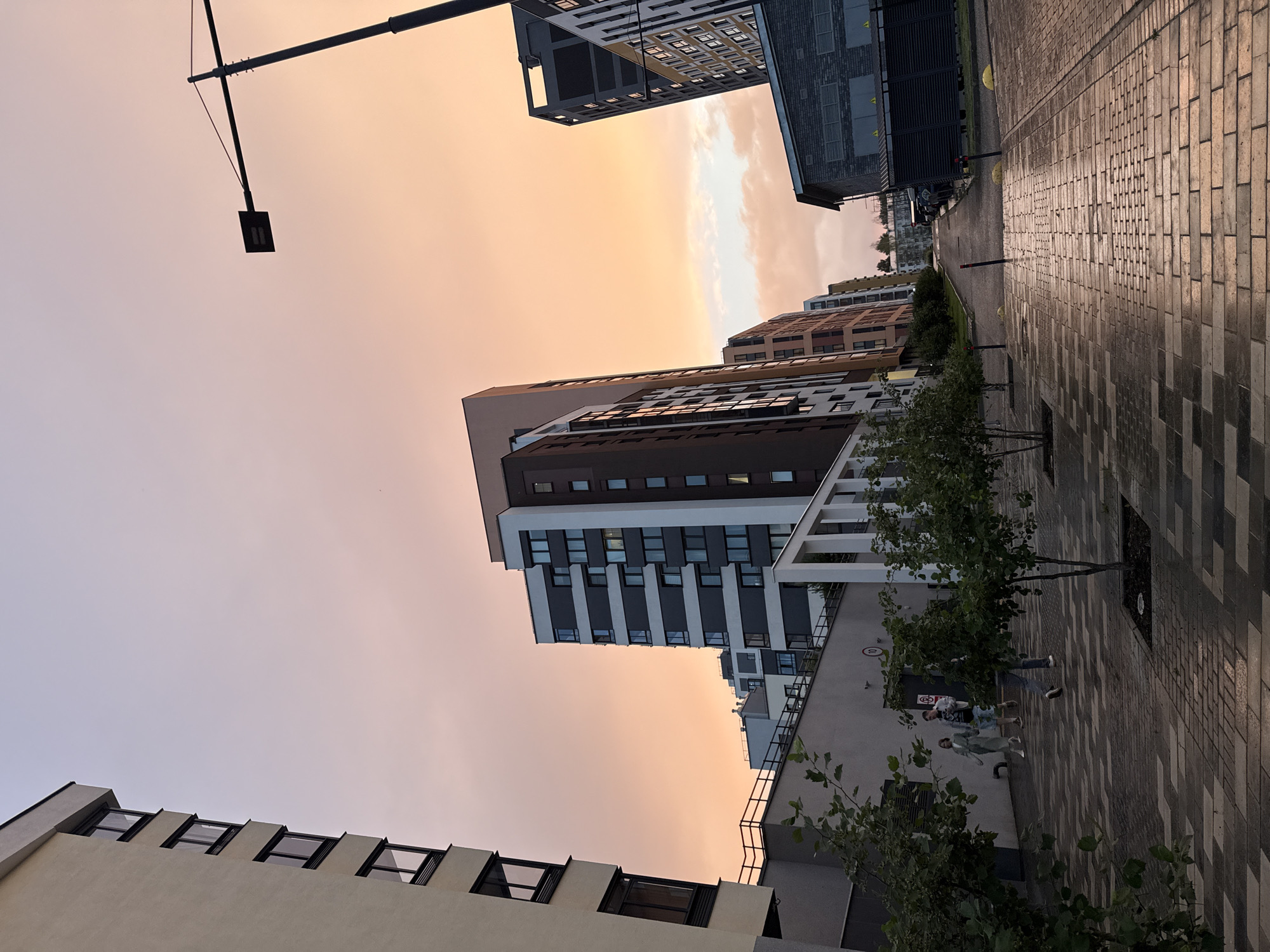 The image captures a street scene in Izhevsk, Russia, characterized by modern residential buildings under a soft, orange-hued sky, indicative of either sunrise or sunset. In the foreground, a paved walkway with a pattern of rectangular bricks is wet, reflecting the ambient light. Small, sparsely leafed trees are planted in square, recessed beds along the walkway.
Several people are walking away from the viewer on the walkway. Two individuals are closer together, with one appearing to be a woman in a light-colored dress and the other a man in a t-shirt and shorts. Further ahead, another person is walking, their legs and lower torso visible.
Dominating the background are multi-story apartment buildings with contemporary architectural designs, featuring a mix of light and dark facades and numerous windows. A street lamp with a rectangular fixture is visible against the sky in the upper right quadrant. The overall environment suggests a newly developed or modern urban residential area. The wet pavement and diffused lighting contribute to a serene, somewhat atmospheric mood.