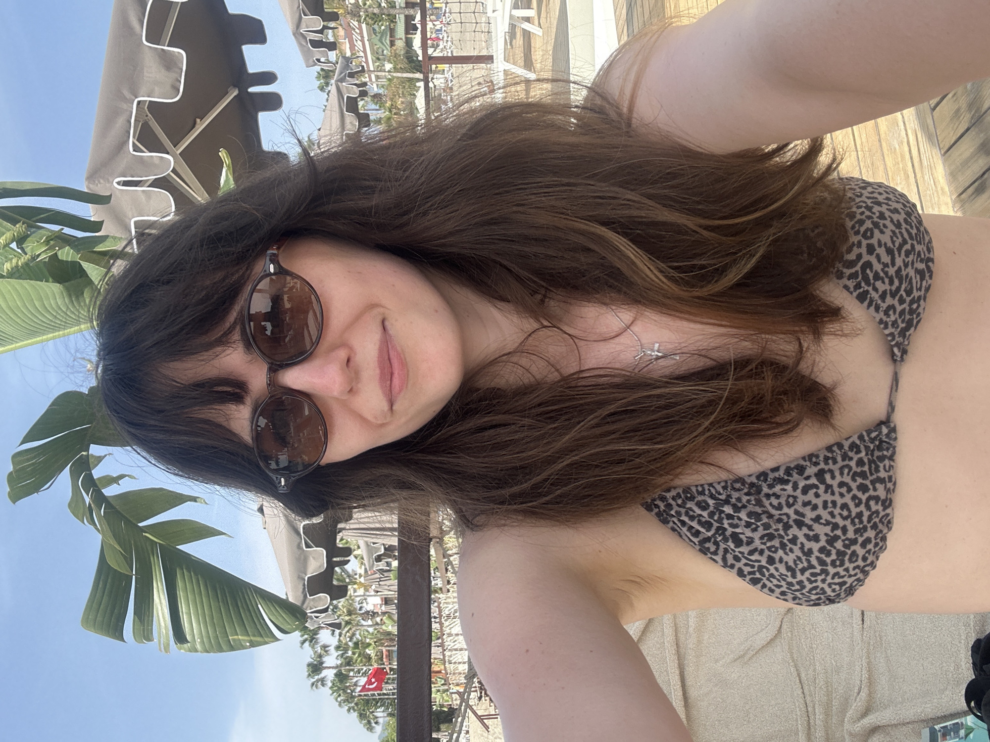 A woman with long brown hair and sunglasses is taking a selfie outdoors. She is wearing a leopard print bikini top. The background shows a beach setting with palm trees, lounge chairs, and beach umbrellas. A Turkish flag is visible in the background. Based on the presence of the flag and beach setting, the location is most likely Alanya, Turkey.