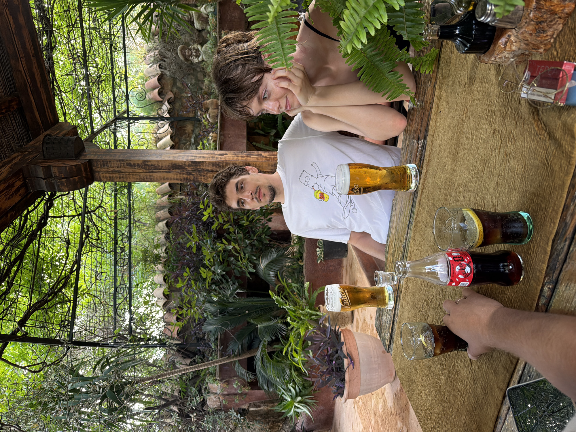 The media file depicts two young adults seated at a table outdoors, surrounded by lush greenery. A man with dark curly hair and a goatee wears a white t-shirt with a cartoon graphic. He is looking towards the camera. Beside him, a young woman with light brown hair is leaning on her hands, her gaze directed downwards with a contemplative expression. She is wearing a black tank top.
On the rustic wooden table, several beverages are visible. There are at least two glasses of golden liquid, appearing to be beer, and several glasses and a bottle of Coca-Cola. A bottle of what seems to be a dark spirit is also present, along with a pair of reading glasses and some small books. The table is covered with a textured, light brown cloth.
The setting appears to be a garden or a patio, with abundant plants and trees, including ferns and palm-like foliage, creating a tropical or Mediterranean atmosphere. A wooden structure, possibly part of a pergola or gazebo, is visible in the background, along with decorative elements like clay pots and stone features. The overall impression is one of a relaxed, casual gathering, possibly a meal or drinks outdoors. The time of day seems to be daytime, with natural light illuminating the scene. The location context is Kamenovo, Montenegro.