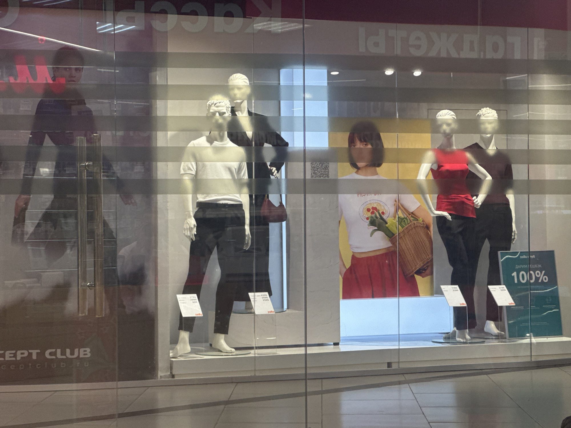 This is a store window display in Ishim, Russia, showcasing mannequins dressed in clothing. The display features several mannequins, some of which are partially obscured by horizontal blinds or reflections. On the left, there is a mannequin dressed in dark clothing, appearing to walk through the window. To its right, two male mannequins are displayed. One is wearing a white t-shirt and black pants, while the other is dressed in a black suit. Further to the right, a female mannequin is shown in a red top and black pants, holding a basket. Next to her is a mannequin wearing a white t-shirt with a graphic design and a red skirt, holding a bouquet of what appear to be vegetables. The background of the display includes posters with text, some of which are in Russian, and a large green sign with "100%" printed on it. There are also price tags visible on some of the mannequins' feet. The overall impression is a commercial display intended to attract customers to a clothing store.