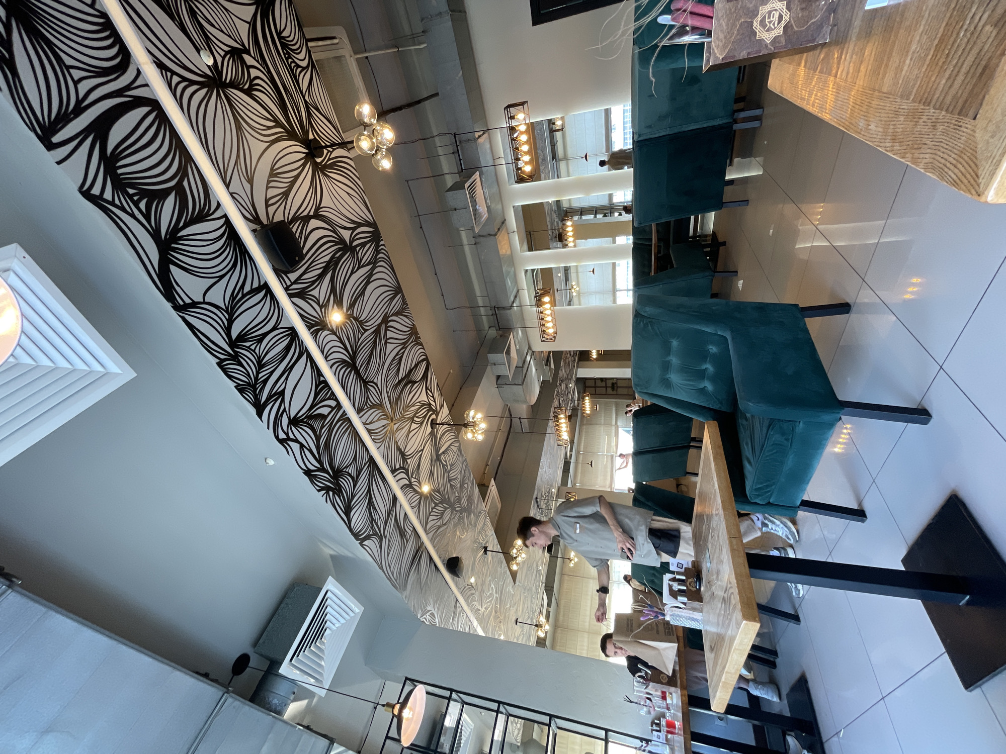 The image depicts an indoor scene, possibly a restaurant or cafe, in Dachnoe, Russia. The interior features a decorative wall with a black and white wave-like design, multiple light fixtures, and rows of teal-colored chairs. Tables with wooden tops are present, and two people are interacting near one of them. One person is standing and appears to be wearing a light-colored shirt, while the other is seated at the table. Various items are visible on the table. The floor appears to be tiled.