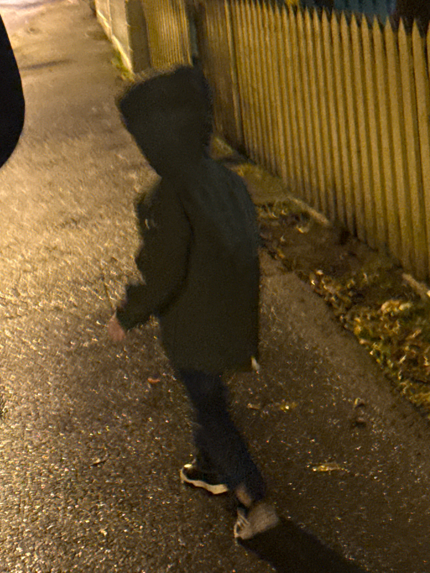 The image shows a child walking on a wet, paved surface next to a wooden fence. The child is wearing a dark hooded jacket and dark pants, with athletic shoes. The fence is made of vertical wooden planks and appears to be part of a property line. The ground is illuminated by artificial light, casting shadows. The scene suggests an outdoor urban or suburban environment in Tallinn, Estonia, possibly a sidewalk or pathway alongside a street. The overall impression is one of a cool, possibly damp, evening.