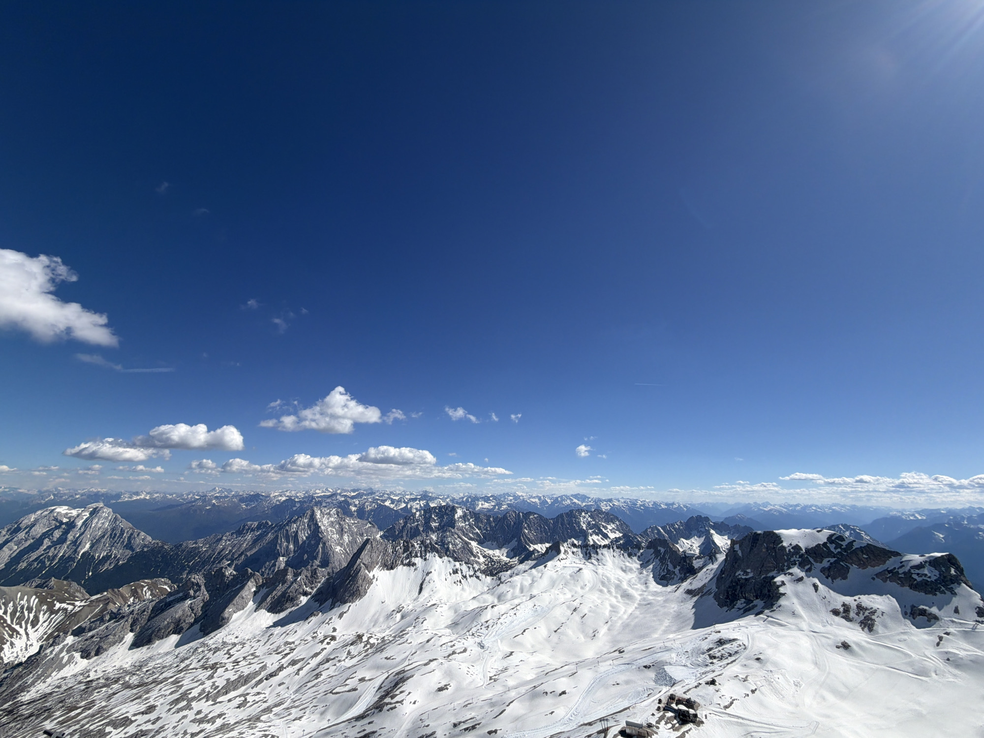 A wide-angle, eye-level outdoor shot shows snow-covered mountain ranges under a clear blue sky with a few scattered clouds. The mountains are rugged and textured with rocky outcrops emerging through the snow cover. In the foreground, there's evidence of tracks in the snow. This appears to be a view from the summit of Zugspitze, Germany's highest peak, located in Grainau.