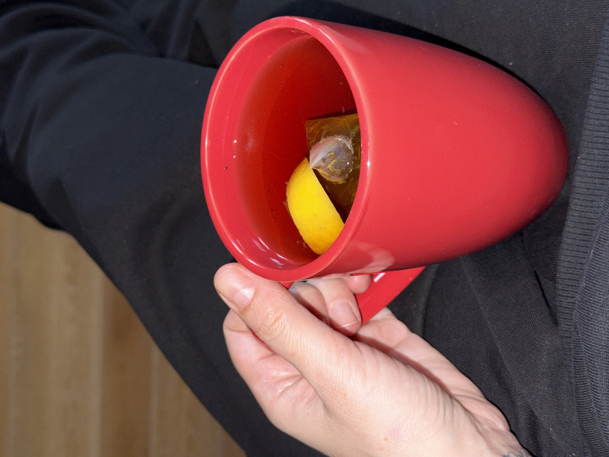 A hand holds a red mug containing a tea bag and a lemon slice. The mug is filled with a light brown liquid, presumably tea. The person wearing black clothing is holding the mug. A wooden surface is visible in the background. There is no evidence indicating this image was taken in Moscow, Russia.