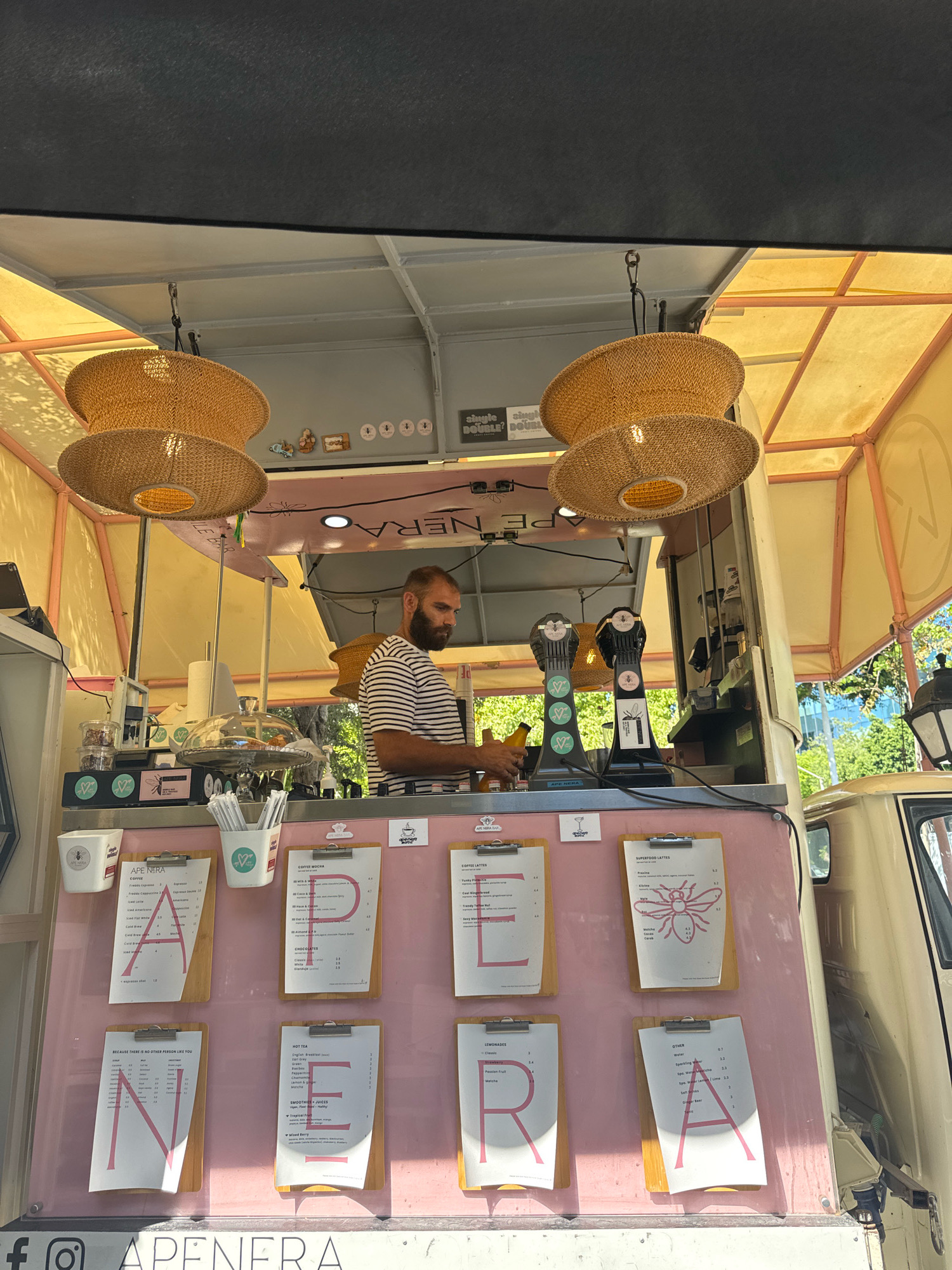 An exterior medium shot captures a mobile coffee shop named "APE NERA" in Limassol, Cyprus. The shop is housed within a van, with its side opened to reveal a counter and a barista. A male barista with a beard, wearing a striped t-shirt, stands behind the counter, preparing drinks. The counter displays a pink facade with menus clipped to boards, spelling out "APE NERA" with large letter cutouts. Various coffee machines and supplies are visible on the counter. Above the counter, two large, woven lampshades hang from the ceiling, casting a warm glow. The background shows trees and buildings, suggesting an urban environment. The overall scene depicts a lively and inviting coffee service operation.