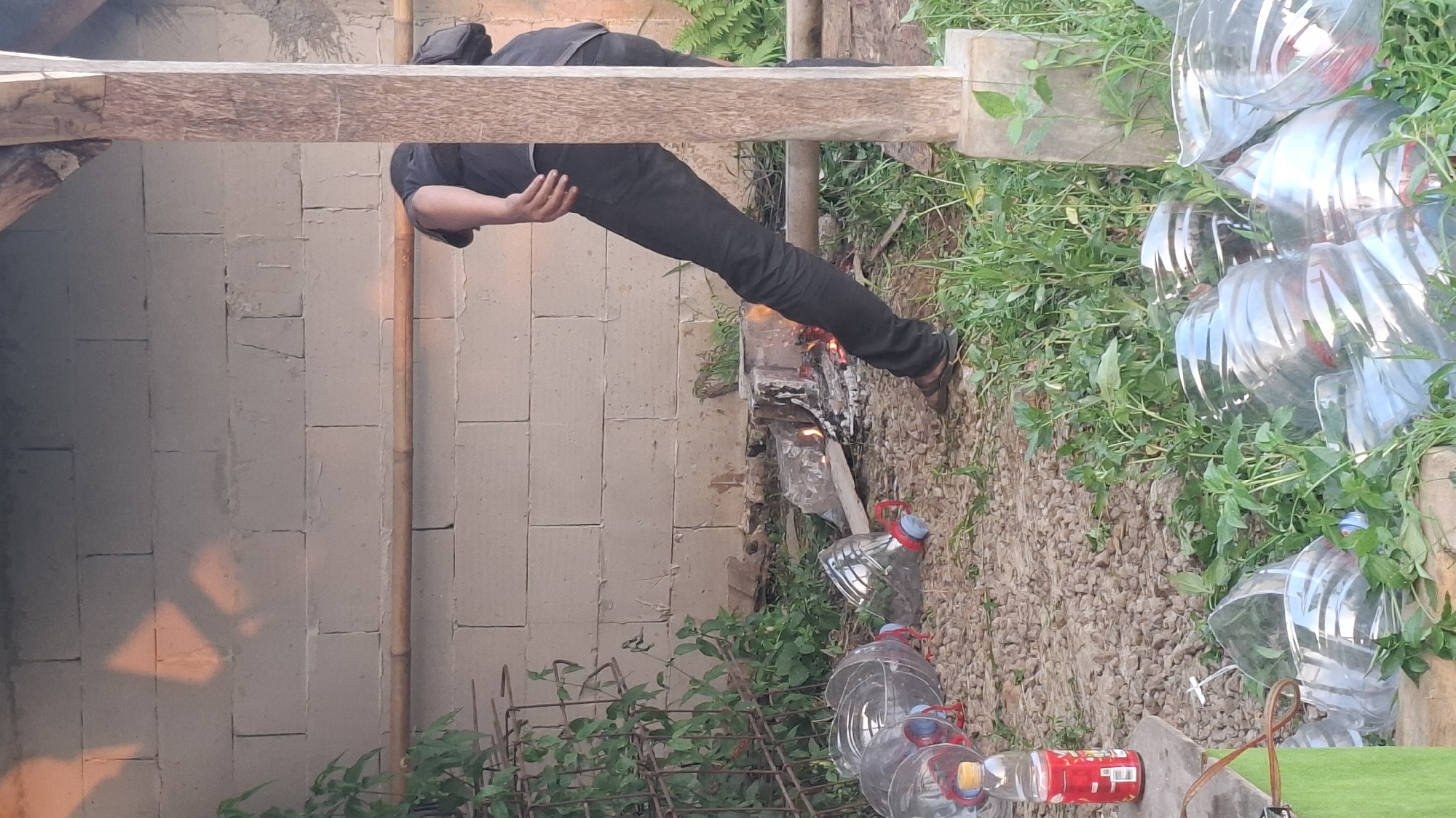 In the city of Rahong, Indonesia, a person, whose head is obscured from view, is bent over a small campfire. The individual is wearing dark pants and a dark shirt, and is leaning on a wooden post. The campfire is built on the ground, with visible flames and burning wood. In the foreground, several large, empty plastic bottles are scattered on the ground, some filled with green foliage. Behind the campfire, a wall constructed from cinder blocks serves as a backdrop. The environment appears to be outdoors, with green plants and gravel covering the ground. A metal structure, possibly a fence or support, is visible on the left side of the image, partially covered by green leaves. The lighting suggests it is daytime, with shadows cast on the cinder block wall.