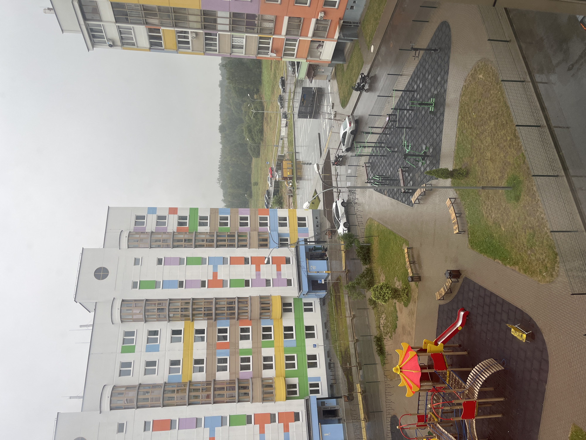 The image depicts a residential complex in Senitsky Selsoviet, Belarus, under an overcast sky. Two multi-story apartment buildings with colorful facades (yellow, orange, pink, purple, green, and blue) are prominently featured. In the foreground, a children's playground with red and yellow equipment, including a slide, is visible. Adjacent to the playground is an outdoor gym with various exercise machines and benches. The ground surfaces consist of paving stones and a rubberized matting in the playground and gym areas. A paved road curves around the complex, with parked cars and motorcycles. In the background, a treeline borders a flat, open area. The scene appears to be during daylight, with wet surfaces suggesting recent rain. There are no people clearly visible in the image.