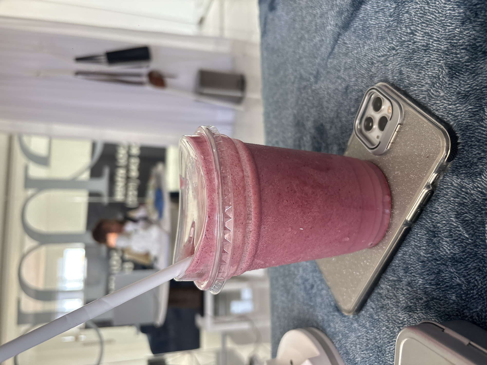 A smoothie in a clear plastic cup with a white straw lies on a phone case, which rests on a blue denim surface. The background includes a partially obscured image of a person in white attire, along with architectural elements. The location is inferred to be Puebla, Mexico, based on the prompt.