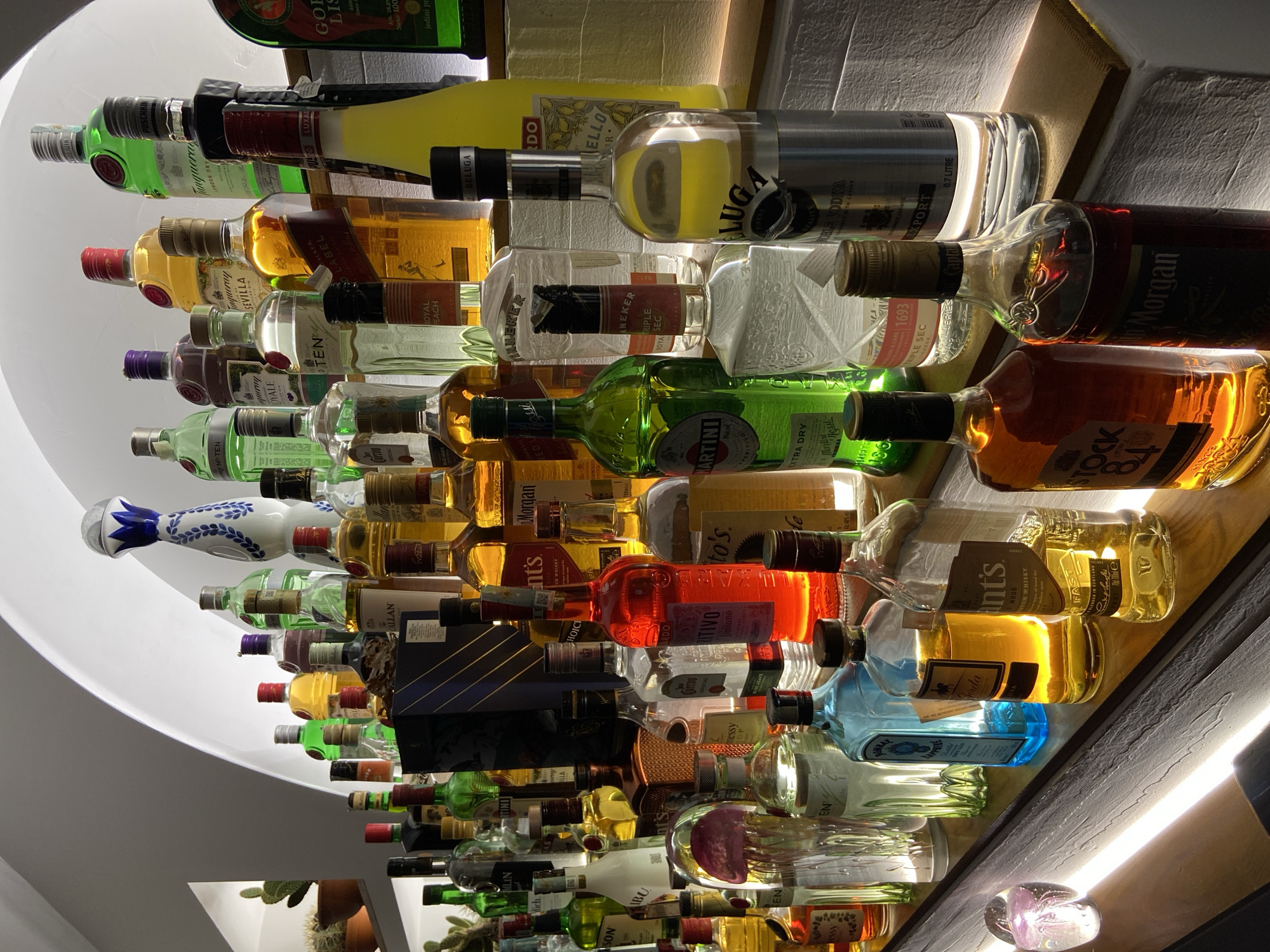 The image shows a well-stocked liquor shelf, likely in a bar or restaurant. The bottles are arranged in rows, with a variety of brands visible, including Tanqueray, Beluga, Martini, Captain Morgan, and Stock 84. The lighting highlights the bottles, creating a visually appealing display. The bottles span different types, from gin and vodka to whisky and liqueur. The shelf is situated against a wall with accent lighting. While the OCR detected the mention of "Petrovac centar, Montenegro", there is no explicit evidence to confirm this as the location within the image itself.
