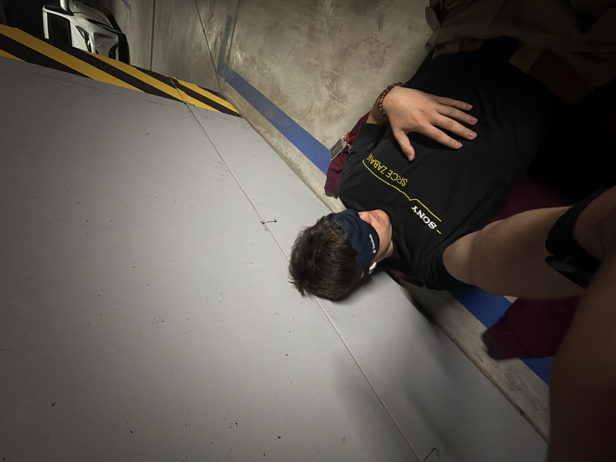 A person wearing a black t-shirt with yellow lettering, a dark blue sleep mask, and white earbuds is lying on their back against a concrete wall in a parking garage. The person has a beaded bracelet on their right wrist. A maroon bag and a keychain with a red tag are on the ground near their head. A white car is parked in the background, and a yellow and black striped pole is visible on the right side of the image. The ground is concrete with blue and yellow markings.