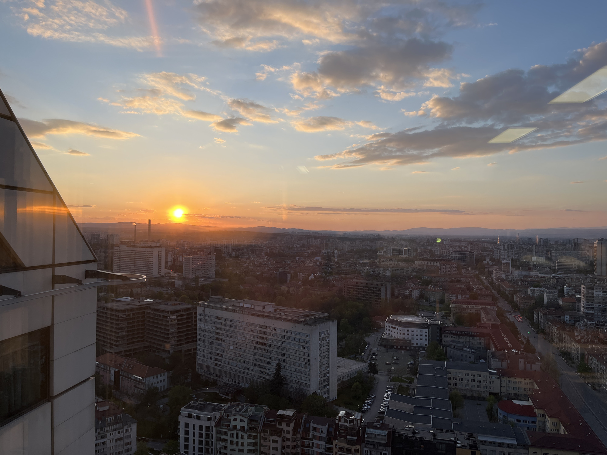 An expansive panoramic view captures the city of Sofia, Bulgaria, at sunset from a high vantage point, likely a tall building with glass walls. The golden sun is prominent on the horizon, casting a warm, soft light across the urban landscape and illuminating scattered clouds in the sky, which transitions from light blue overhead to vibrant oranges and yellows near the setting sun. Distant mountains are visible in the background, silhouetted by the sunset glow.
In the foreground to the left, part of a modern glass-clad building, from which the photograph was taken, is visible, showing reflections of the setting sun and internal structures. Reflections of rectangular interior lights are also visible on the window glass in the upper right portion of the image. The city below is densely packed with a mix of buildings, including several large, multi-story residential or office blocks, some appearing older with concrete facades, and others newer. One partially constructed or dilapidated building clearly displays the Cyrillic text "ХРОМЕ" (CHROME) on its side.
Numerous roads crisscross the urban fabric, with cars visible, some parked and others moving, indicated by small green lights that suggest traffic flow. Green spaces and trees are interspersed among the buildings. The overall scene depicts a calm evening in a bustling city as day turns to night, with no specific human activity discernible beyond the implied ongoing life of the city.