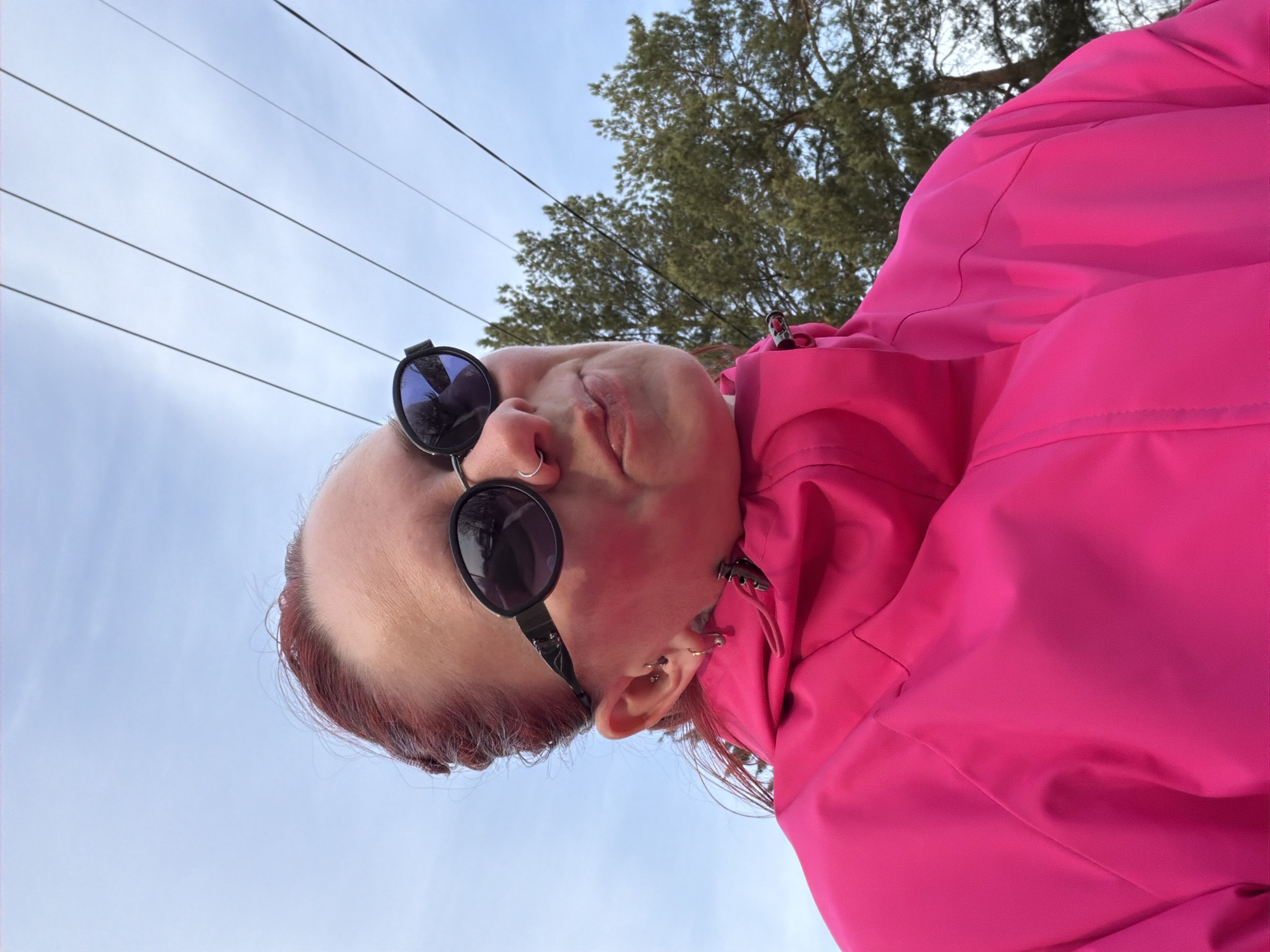 A low-angle shot features a person wearing dark sunglasses and a bright pink jacket. The person is facing upwards, with a blue sky and electrical wires visible in the background. Behind the person's head, there are bare tree branches. The person has multiple piercings in their ear and a nose ring. The location is most likely outside in the city of Chaikovsky, Russia.