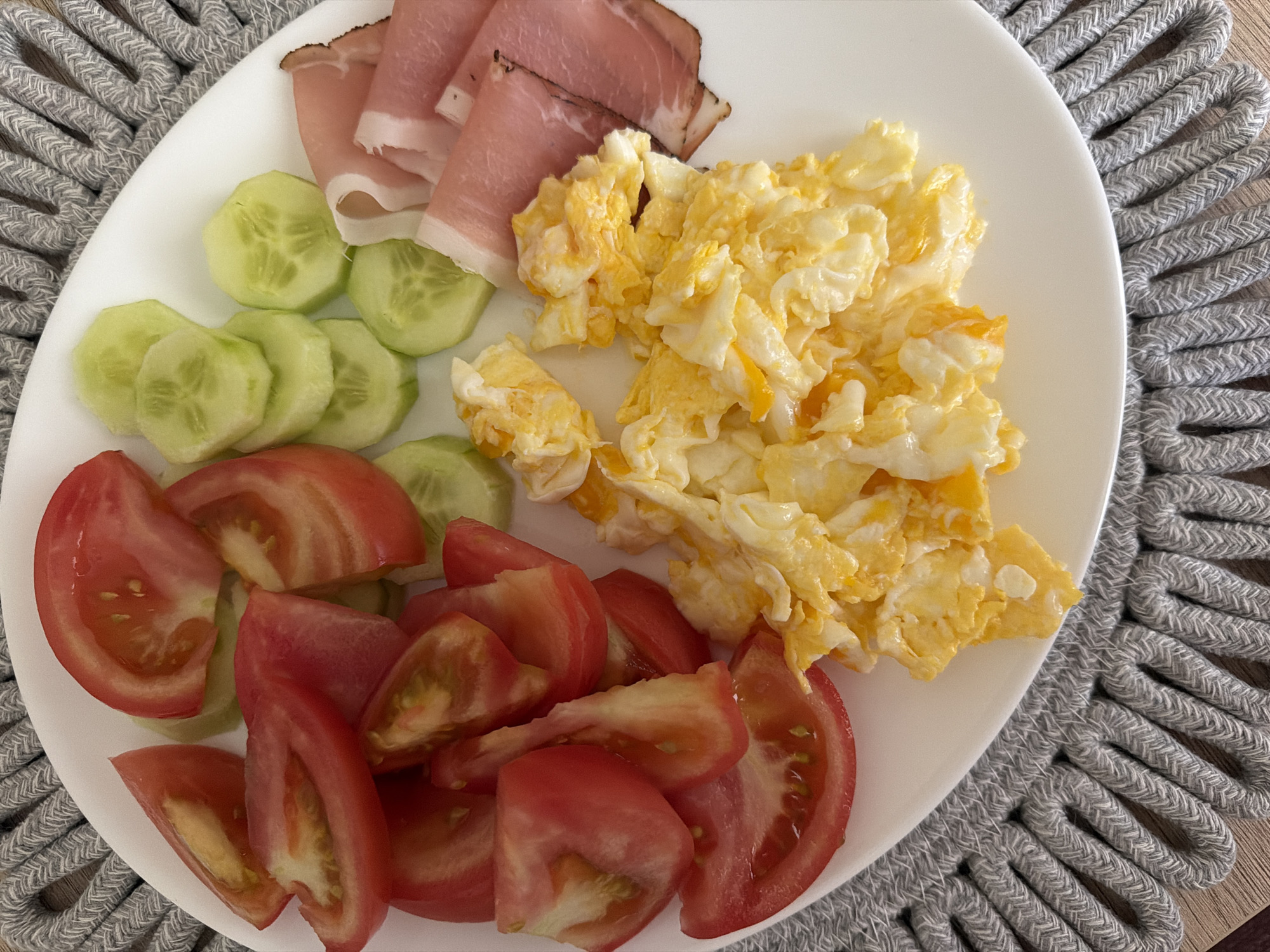 A top-down shot shows a white plate with a breakfast of scrambled eggs, sliced tomatoes, sliced cucumbers, and folded slices of ham. The plate rests on a grey, woven placemat. The background is a wooden surface.
