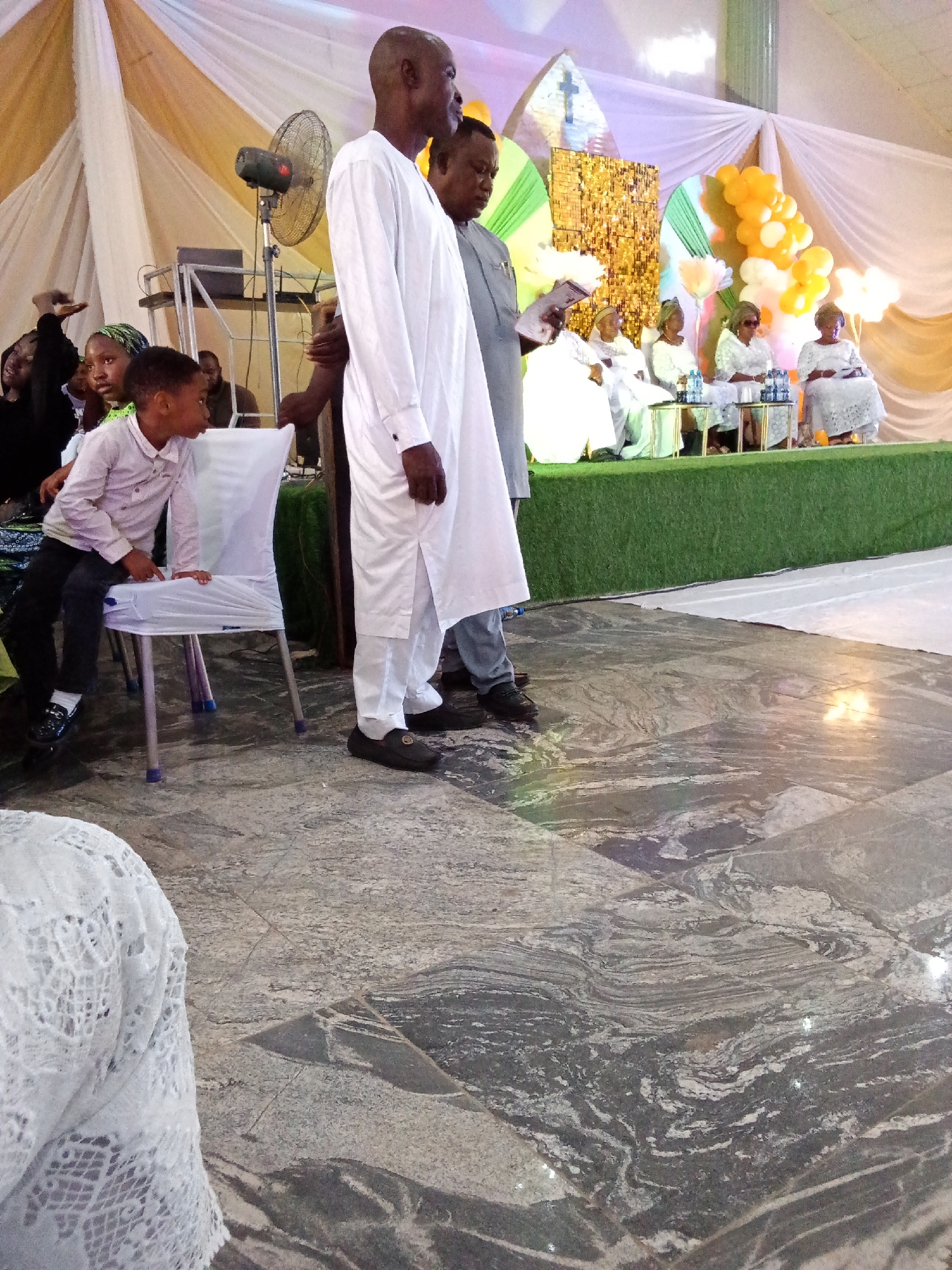 The image depicts an indoor event, possibly a ceremony or gathering, taking place in Barden Yana, Nigeria. In the foreground, a man in a white traditional outfit stands with his side profile visible. To his left, a young boy in a light-colored shirt and dark trousers sits on a white-covered chair. Several other individuals are seated further back on a raised platform covered with green artificial turf. Behind them, the background features draped white and gold fabric, balloons, and decorative elements, including a cross symbol. The floor is a polished stone with a marbled pattern. A floor fan is visible on the left side of the image, near the seated individuals. The lighting suggests an indoor setting, with warm illumination.