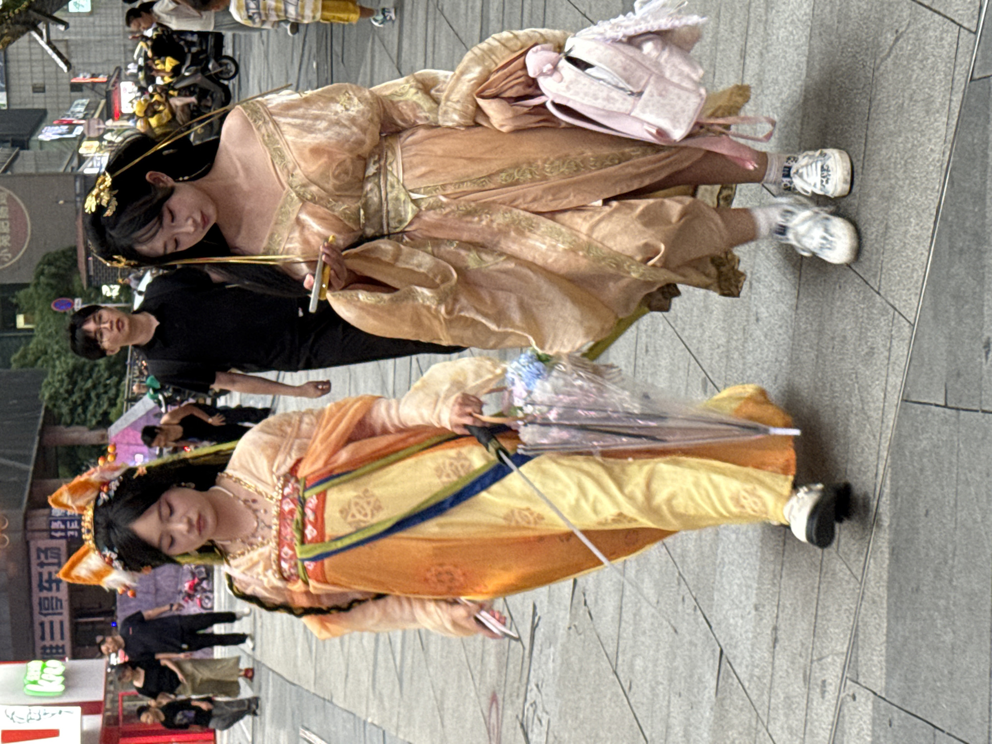 The image depicts two young women dressed in elaborate, traditional Chinese-inspired attire walking on a paved city sidewalk in Yuzhong District, China. The woman on the left wears a vibrant orange and yellow layered garment adorned with intricate patterns, complemented by a beaded necklace and fox-ear hair accessories. She holds a transparent umbrella decorated with blue flowers in her right hand and a silver object in her left. The woman on the right is dressed in a flowing, pale peach-colored gown with gold embroidery, featuring an off-the-shoulder design and ornate gold hair accessories. She is looking at a mobile phone in her right hand and carries a pink backpack adorned with white ribbon details on her right side. Several other individuals and a motorcycle are visible in the background, suggesting a busy urban environment. The sidewalk is made of grey stone tiles with visible grout lines, and storefronts with signage are present on the left side of the frame.