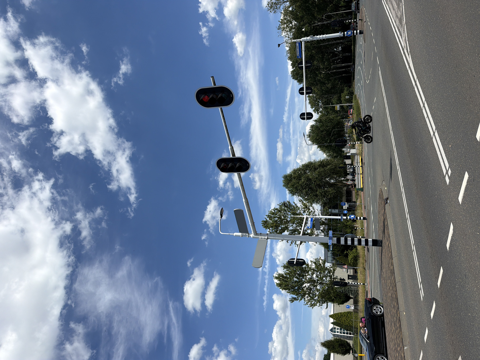 The image shows a traffic intersection in Helmond, Netherlands, under a bright blue sky with scattered white clouds. In the foreground, a black convertible is driving away from the camera, with two passengers visible. To the right, a motorcyclist is riding through the intersection. Traffic lights are visible, with some indicating red. There are trees and buildings in the background, suggesting an urban environment. The road is asphalt with white lane markings. The overall scene captures a moment of everyday life at a Dutch intersection on a sunny day.