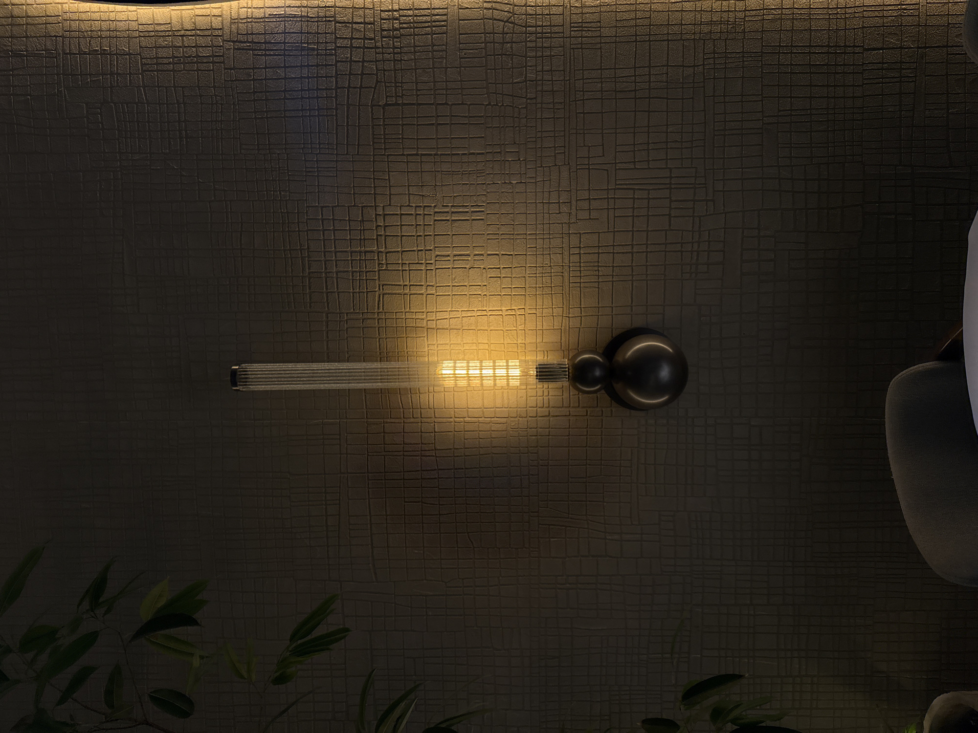 The image displays a dimly lit indoor setting, focusing on a dark, textured wall. A prominent wall-mounted light fixture, or sconce, is positioned horizontally across the center of the frame. This fixture features a long, linear bulb emitting a warm, yellowish light, and two spherical, dark metallic elements on its right side.
The wall itself is the most striking subject, covered in an intricate, irregular grid pattern that appears to be embossed or deeply textured, possibly resembling a stylized woven fabric or alligator skin. The warm light from the fixture illuminates this texture, highlighting the relief and creating subtle shadows that enhance its depth. A softer, indirect light source also illuminates the top portion of the wall, creating a glowing effect along the ceiling line.
In the bottom-left corner, the vibrant green leaves of a plant are visible, providing a natural contrast to the predominantly dark and artificial elements. To the right side of the image, a partial view of a dark, possibly upholstered, piece of furniture, such as a chair or sofa, can be seen.
The scene conveys a sophisticated and intimate atmosphere, indicative of an evening or night setting due to the reliance on artificial lighting. No people or specific activities are depicted. No text is visible in the image. While the location context of Yerevan, Armenia, is provided, there are no discernible visual cues within the photograph to specify this geographical location.