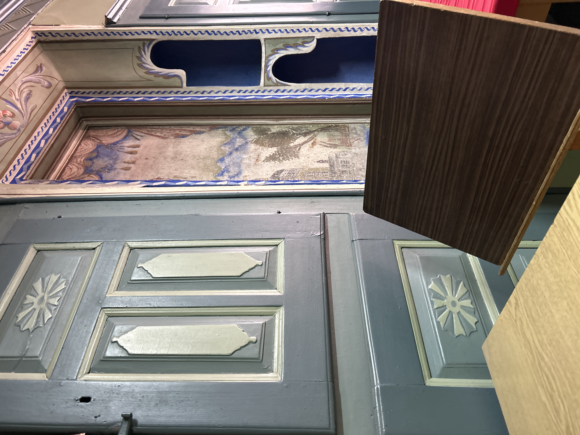 The image shows an ornately decorated interior, likely a historical building in Plovdiv, Bulgaria. The ceiling is painted with intricate designs and a scene, possibly depicting a landscape or cityscape. Below the ceiling, a blue-grey door or panel features decorative mouldings and carved flower-like motifs. Two pieces of wood, one dark and one light, are partially obscuring the view in the foreground. The overall impression is that of a well-preserved or restored historical space with elaborate craftsmanship.