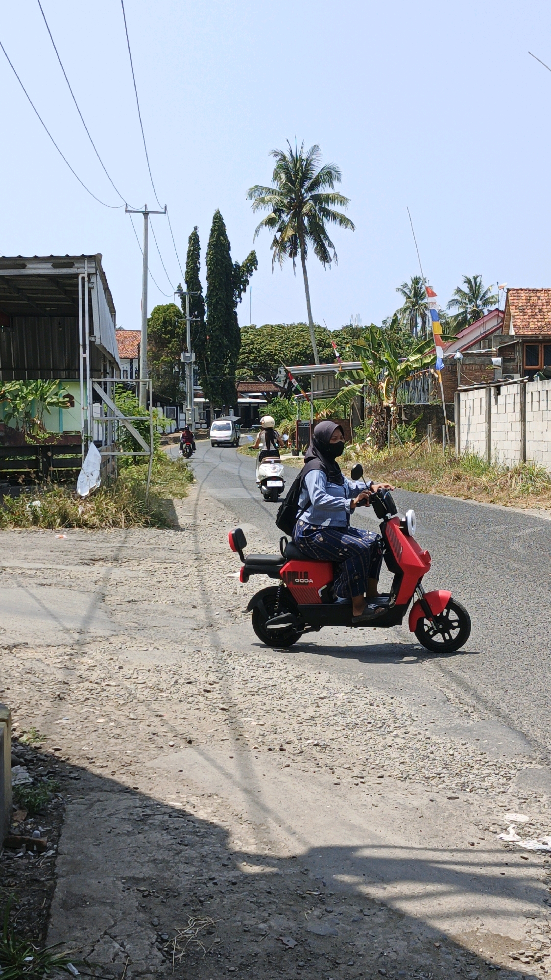 In Desa Karangjaladri, Indonesia, on a sunny day, a woman wearing a black hijab and a blue shirt rides a red electric scooter down a paved road. She is also wearing a black face mask and patterned pants. The scooter has the word "GOODA" written on its side. Behind her, other individuals on scooters and a white car are visible on the road. The environment is characterized by scattered houses with red-tiled roofs, lush green trees including palm trees, and utility poles with visible wires. The road is lined with gravel and concrete on the sides, with some vegetation growing. The scene depicts a typical day in a semi-rural Indonesian village.