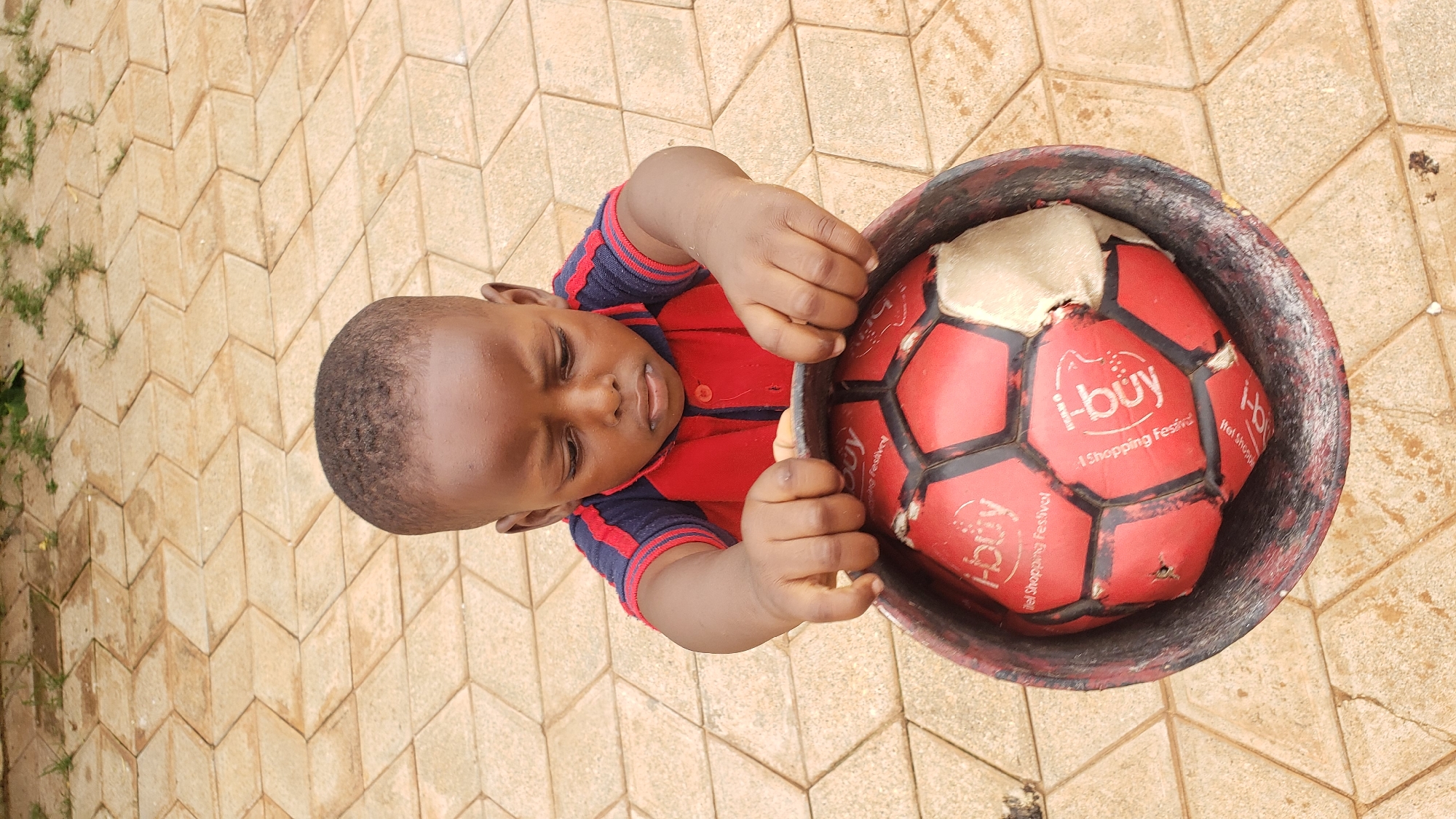 A young boy, wearing a red and blue striped shirt, is holding a red soccer ball in a dark-colored bucket. The ball is partially deflated and has white stitching. The boy is looking down at the ball with a neutral expression. The scene appears to be outdoors, with a tiled ground in the background. Some blades of grass are visible between the tiles. The lighting suggests it is daytime. Text on the ball is visible, including "i-buy" and "Shopping Festival". The overall impression is of a child engaged with a toy in a casual setting.