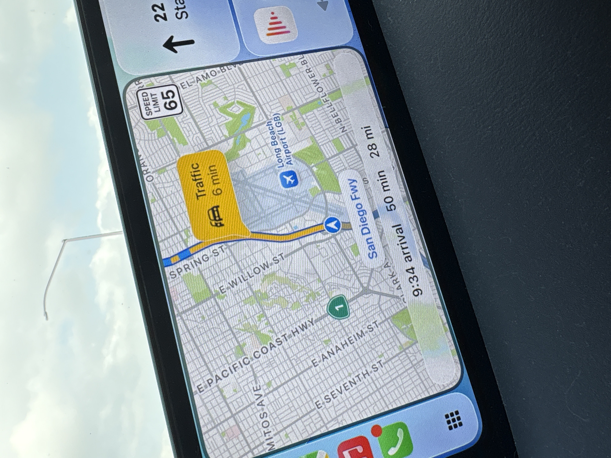 The image shows a map on a smartphone display, displaying navigation information in the city of Long Beach, United States. The map indicates "Traffic 6 min" on a section of a road near Spring Street, which runs to the North of Long Beach Airport. The airport itself is marked with a blue airplane icon. The route is also indicated on the San Diego Freeway. The speed limit is 65. The time of arrival is 9:34, with a remaining drive time of 50 minutes for 28 miles. Roads like E Pacific Coast Highway, E Anaheim St, and E Seventh St are also labeled on the map. The background suggests the photo was taken inside a vehicle.