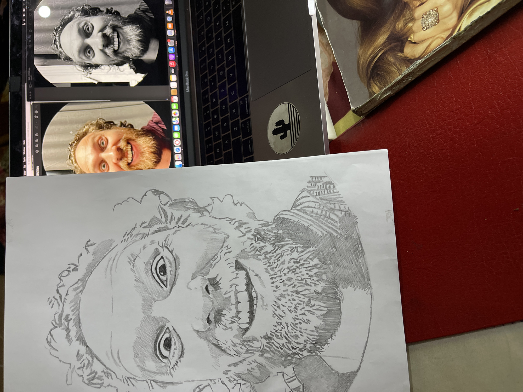 Here is a detailed description of the image:
The image depicts an indoor scene featuring various items arranged on a surface, likely a desk or table. The focus is on a hand-drawn pencil sketch of a man's face, placed on top of a partially visible Macbook Pro laptop. The screen of the laptop displays two circular images of the same man with a beard, one in color and the other in black and white. A sticker depicting a cactus is affixed to the laptop's surface near the trackpad.
To the upper right of the laptop is a magazine or book with a picture of a woman wearing a large diamond ring. A red surface, possibly a desk or tablecloth, occupies the right side of the frame.
The scene lacks information that could pinpoint the location in Jos, Nigeria.