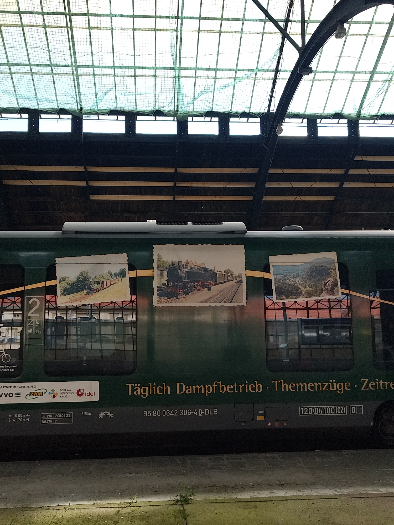 The image depicts a green train car parked at a station in an indeterminate city. The train car features images reminiscent of old photographs pasted onto its exterior, showcasing historical scenes of trains and landscapes. Text on the car reads "Täglich Dampfbetrieb. Themenzüge. Zeitre," suggesting a daily steam operation or themed train journey.
Above the train, the station's architecture is visible, with an intricate framework of beams and a grid-like ceiling structure. The scene is illuminated by natural light filtering through the station's roof.
The overall impression is that of a vintage or heritage train presentation, possibly at a railway museum or a station offering historical train rides.