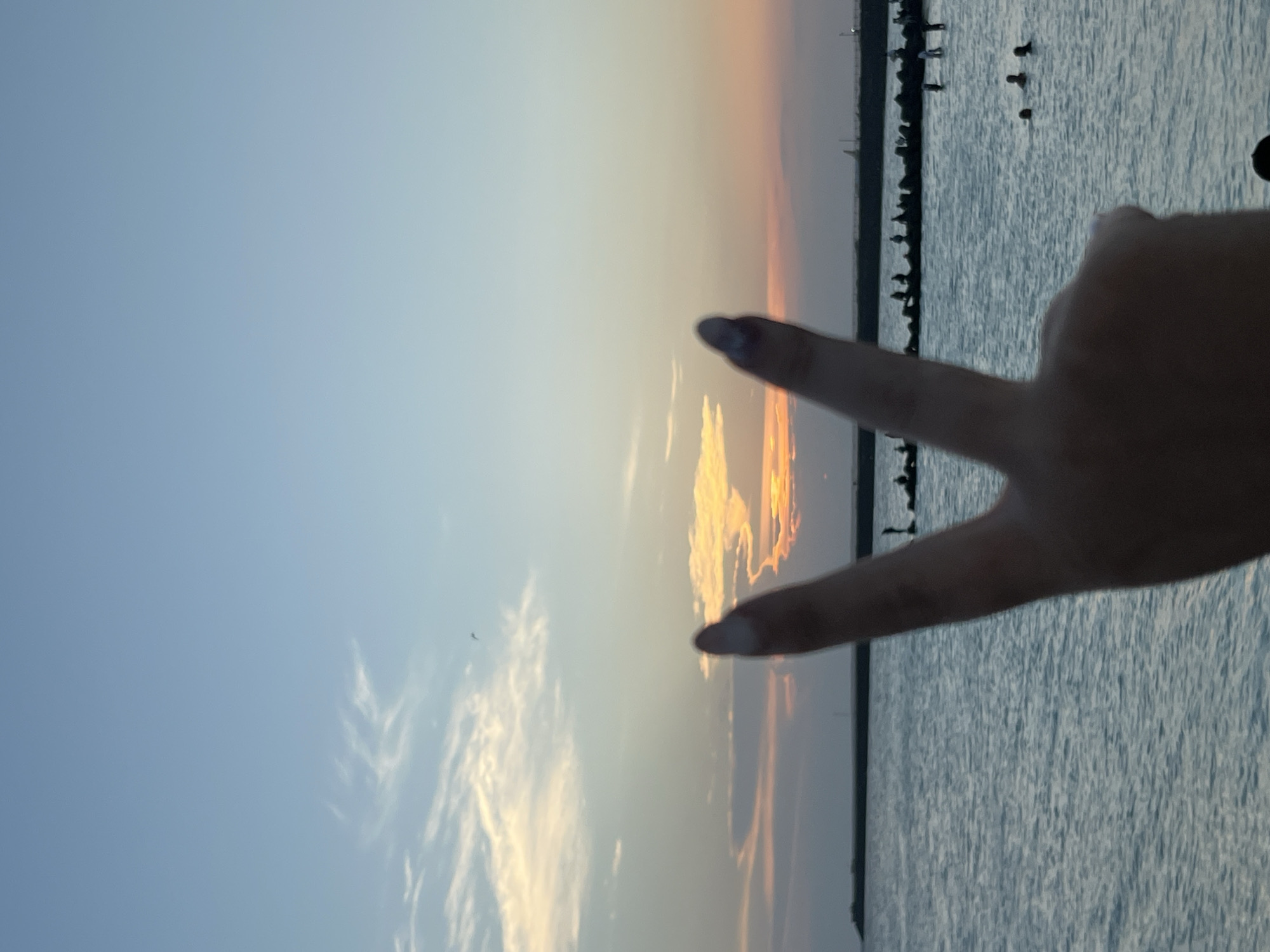 A hand is held up in the foreground, displaying a peace sign. The fingers are blurred, obscuring their details. Behind the hand, a body of water stretches out, with gentle waves rippling across its surface. In the distance, a line of dark, rocky structures runs horizontally, with small silhouetted figures standing or sitting on them. Further out, a few people are visible in the water, swimming. The horizon is marked by a faint line, beyond which the sky transitions from a pale blue at the top to hues of orange and yellow near the horizon, indicating a sunset. Wispy clouds are scattered across the sky. The overall scene suggests a beach or coastal area at dusk.