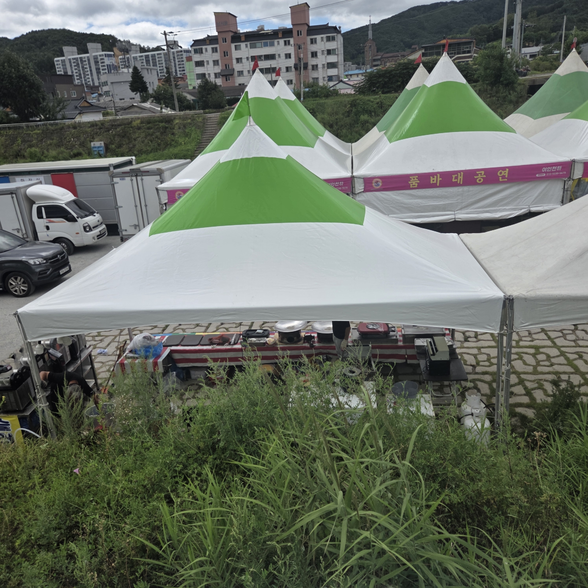 The image shows an outdoor event with multiple white and green striped tents set up on a paved area. In the background, there are apartment buildings and hills. In the foreground, there is lush green vegetation. Beneath one of the tents, there is a table set up with what appears to be food preparation equipment, including pots and a portable stove. A person is visible behind the table, seemingly engaged in cooking or serving. Several trucks are parked in the vicinity, suggesting a mobile market or festival. The location is identified as Dunnae-myeon, South Korea.