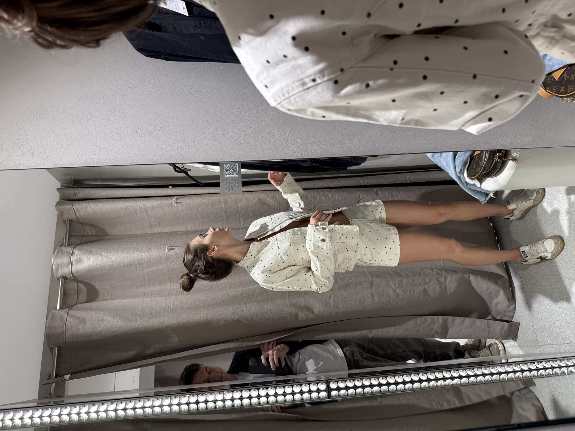 A young woman is standing in a retail changing room, as seen through a mirrored surface. She is wearing a white, short-sleeved, polka-dotted shirt and matching shorts, and white athletic shoes. A man is visible in the mirror's reflection, lying on the floor and holding a cell phone. The changing room has gray curtains, and a clothing tag with a QR code is partially visible above the woman. The location is described as Unknown, Unknown.