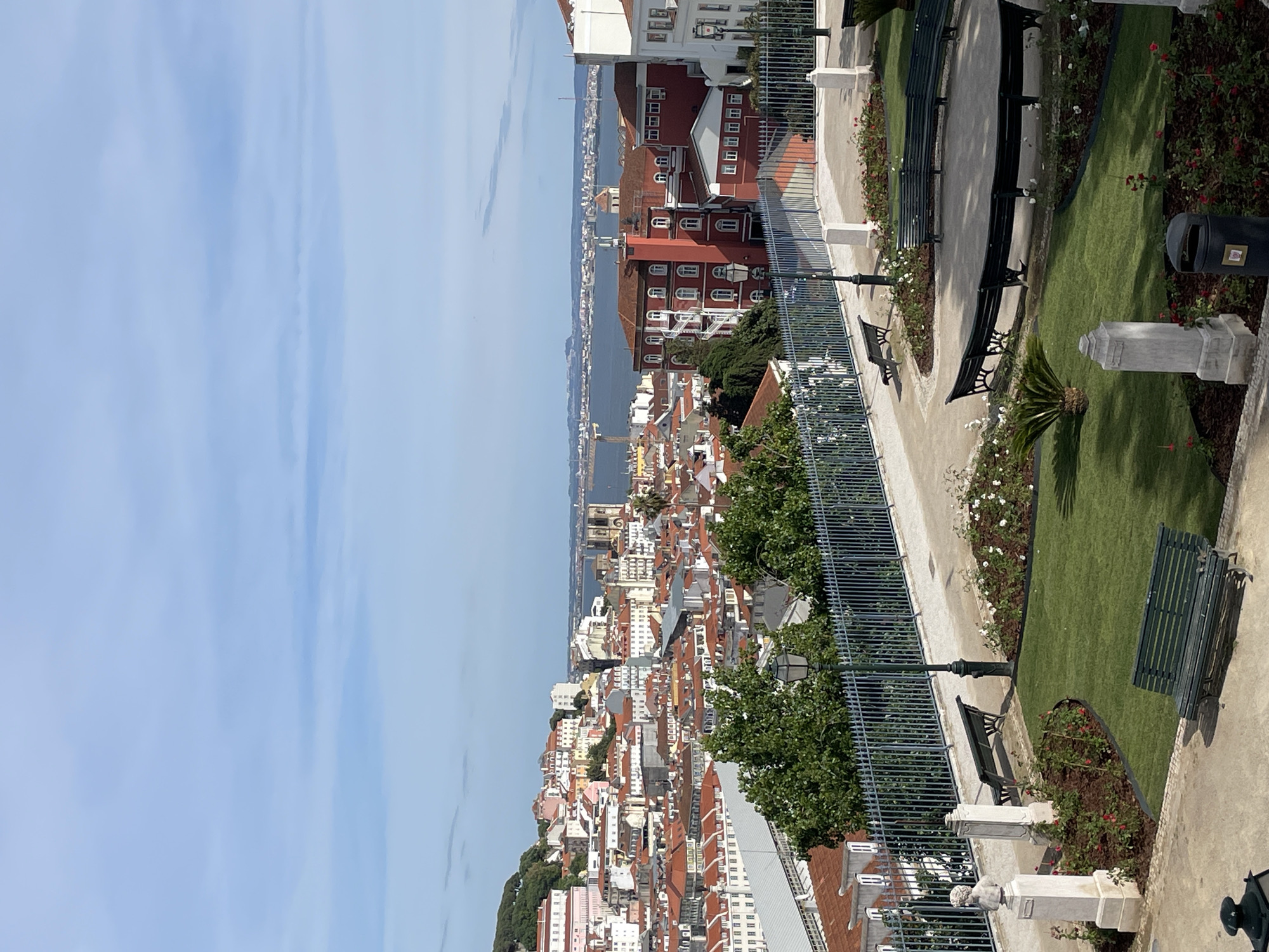 The image presents an elevated view of Lisbon, Portugal, likely taken from a viewpoint or garden within the city. Below, a dense urban landscape unfolds, characterized by buildings with predominantly red-tiled roofs. A body of water extends into the distance, suggesting a river or the sea. In the foreground, there is a park area with benches and flower beds, typical of a maintained urban garden. A metal fence separates the garden from the city view. No people are directly visible in the frame, but the presence of benches implies a public space intended for relaxation and viewing the city. The lighting indicates a sunny day with clear skies.