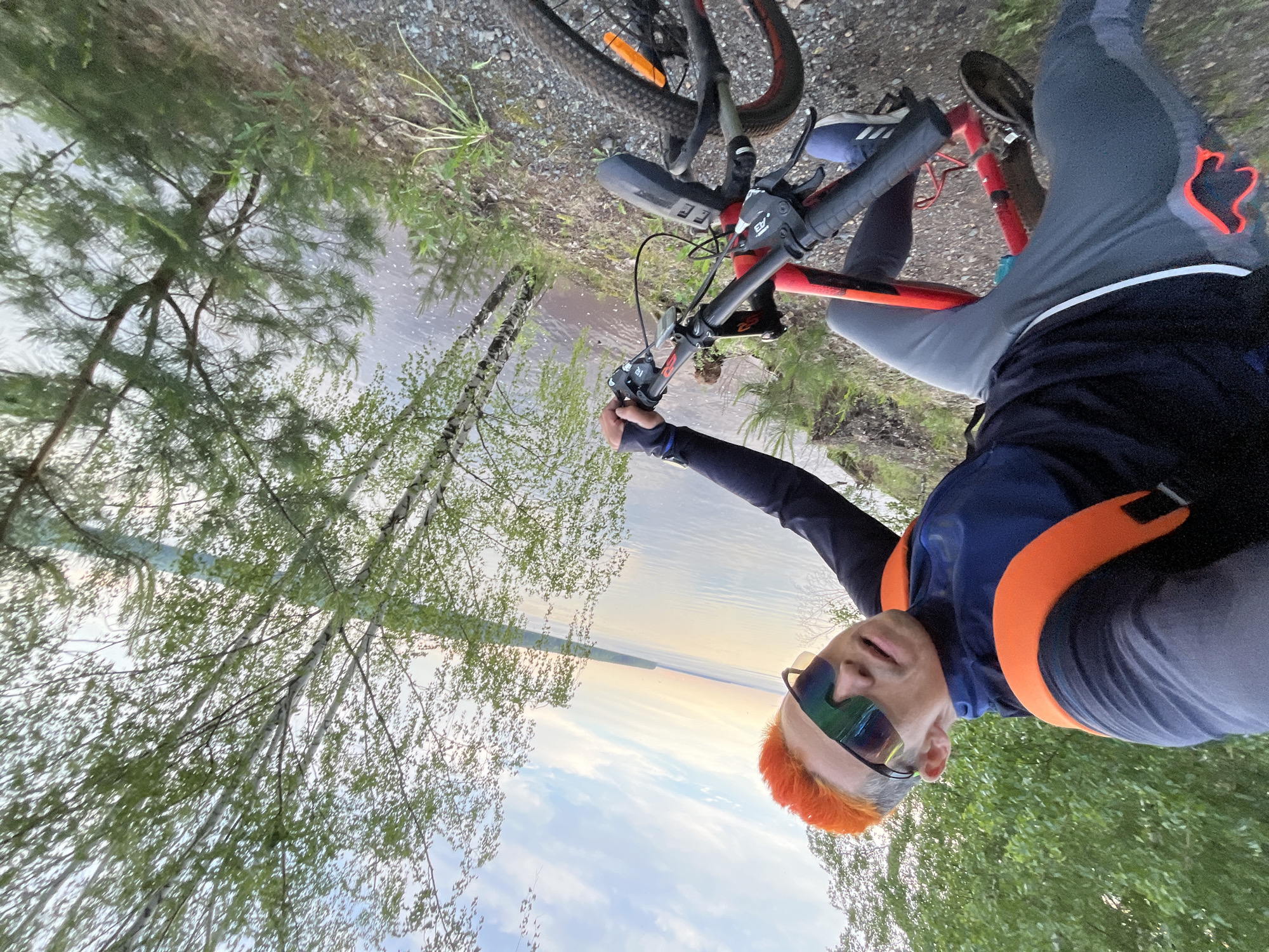 A person is taking a selfie while holding onto the handlebars of a red and black mountain bike. The background includes a body of water and trees. The person has orange hair and is wearing sunglasses, a dark blue and orange vest, and gray athletic pants. The location is likely in the city of Chaikovsky, Russia, based on the prompt. The ground appears to be a gravel path near the edge of the water. The photo is taken at an angle.